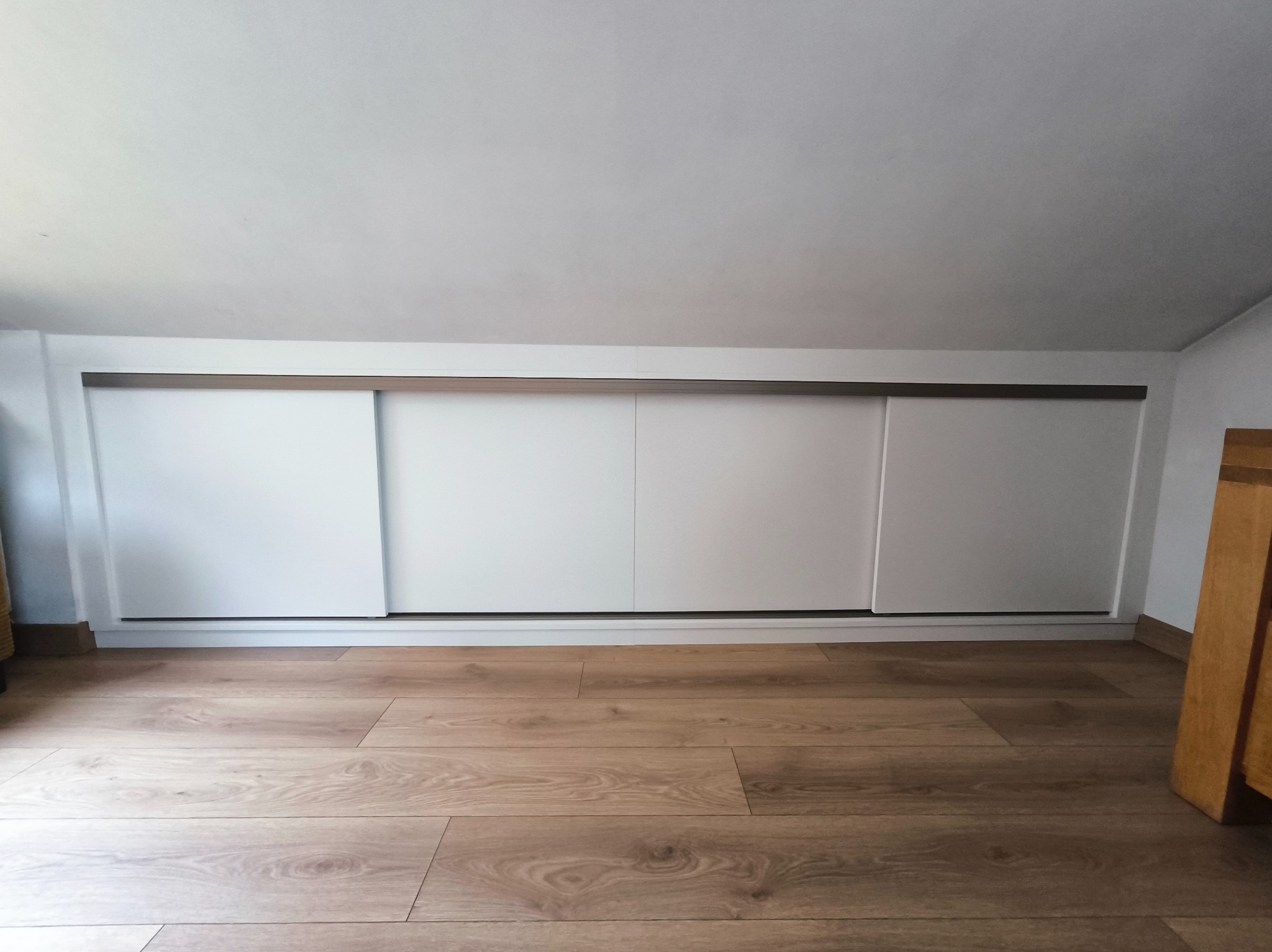 Biała szafa wnękowa z przesuwnymi drzwiami, zamontowana pod skosem dachu na poddaszu, z drewnianą podłogą. Minimalistyczny design.