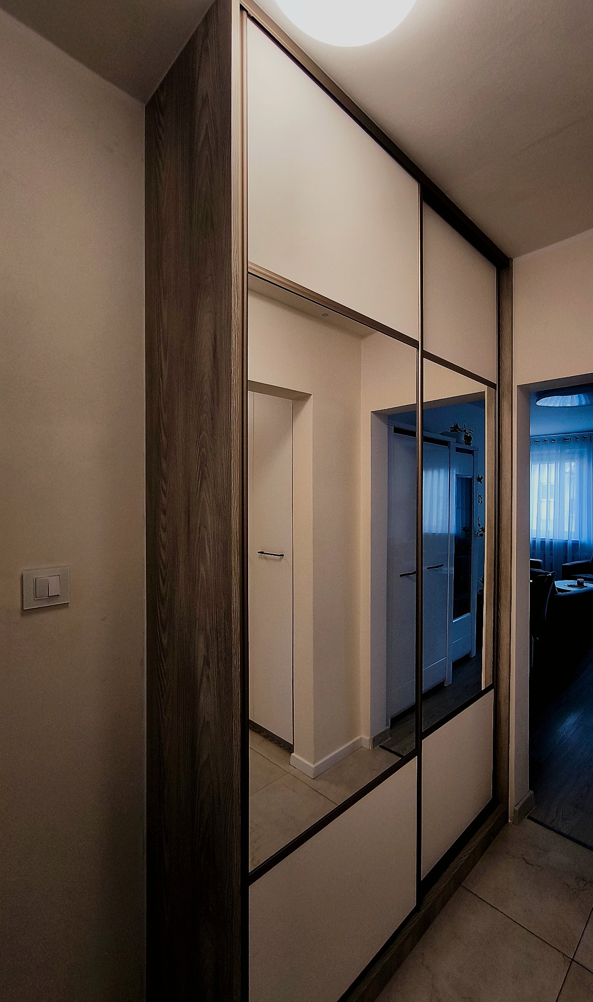 Nowoczesna szafa wnękowa z przesuwnymi, lustrzanymi drzwiami, z elementami w kolorze drewna, w przedpokoju. Widoczne odbicia wnętrza mieszkania.