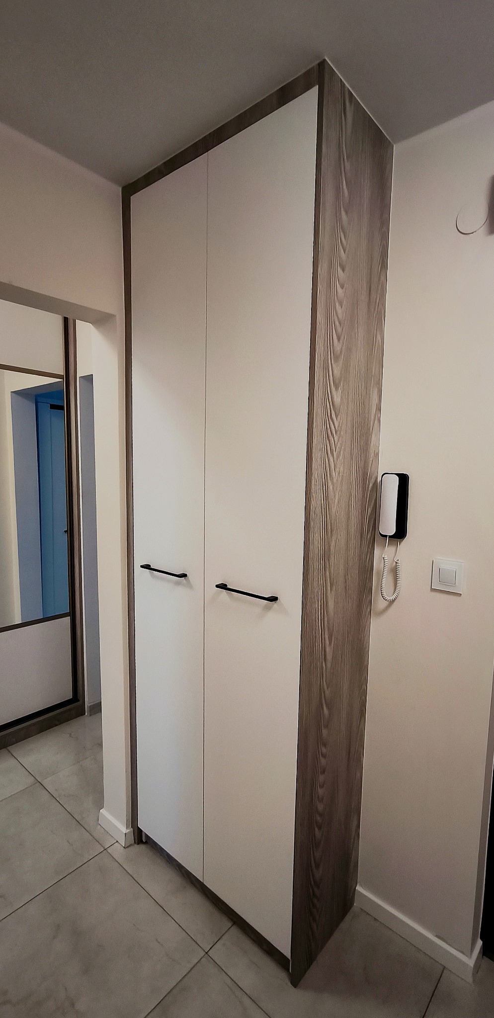 Dwudrzwiowa szafa na wymiar z białymi frontami i drewnopodobnym bokiem w przedpokoju. Widoczne czarne, podłużne uchwyty i fragment lustrzanej szafy przesuwnej.