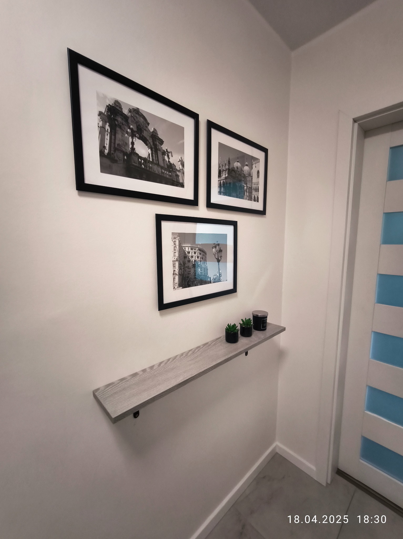 Zamontowana półka ścienna z dekoracjami, pod trzema czarno-białymi obrazami w ramkach. Widoczny fragment drzwi z niebieskimi akcentami.