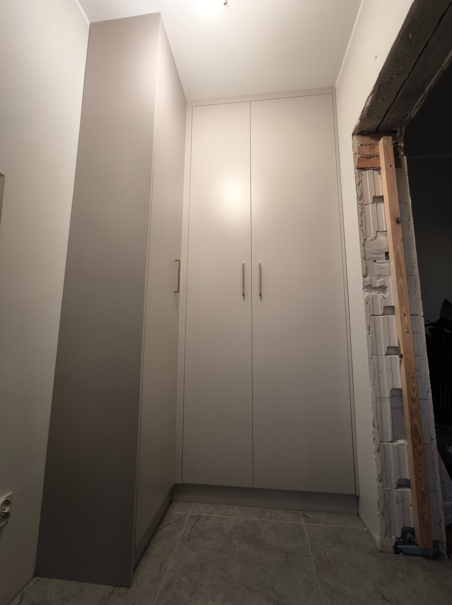 Jasnoszara szafa wnękowa z minimalistycznymi uchwytami, dopasowana do narożnika, widoczna obok surowego otworu drzwiowego w trakcie remontu.