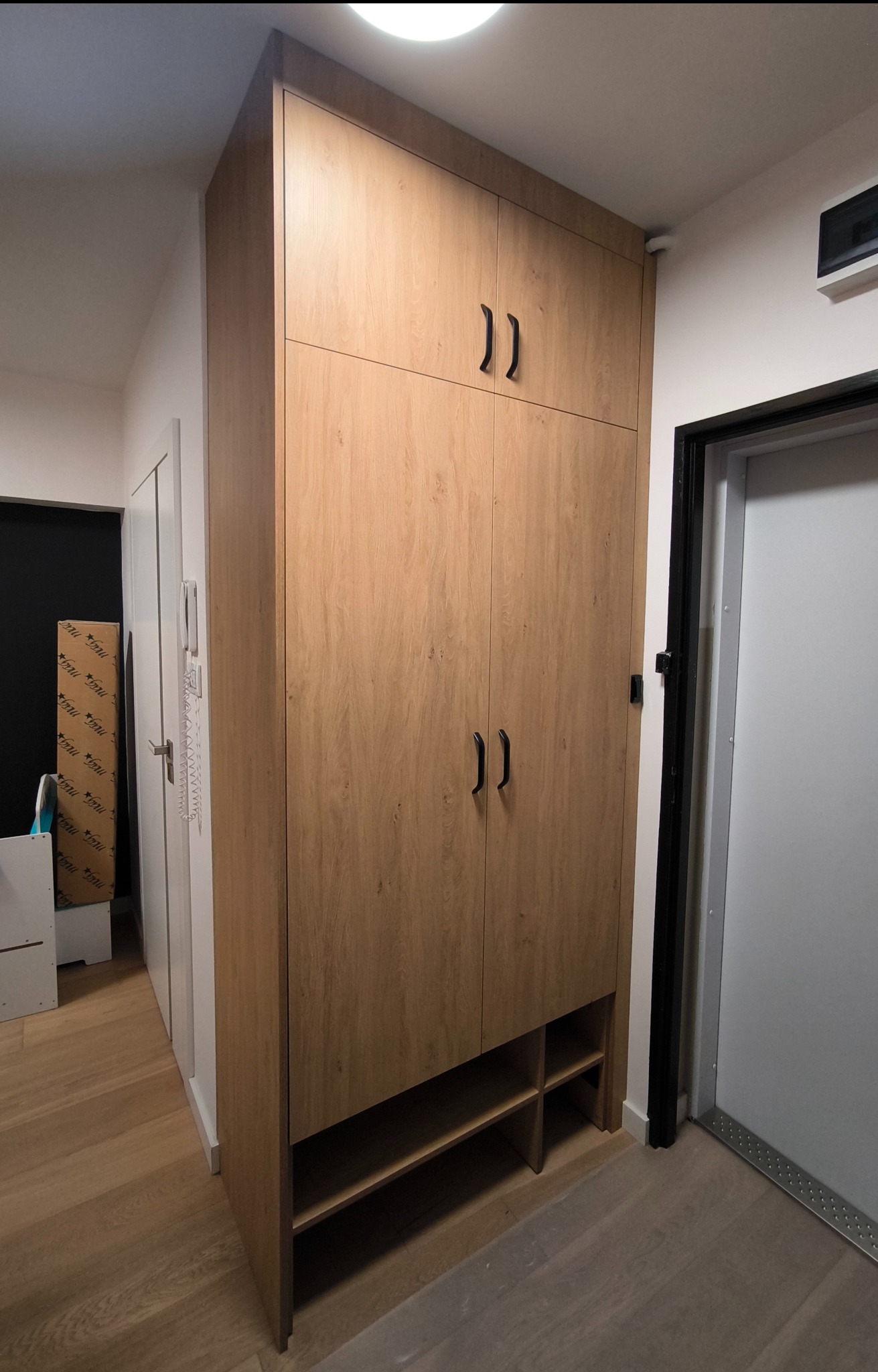 Pełna szafa wnękowa z drewnopodobnymi frontami i czarnymi uchwytami, z otwartymi półkami na dole, wkomponowana w przestrzeń korytarza przy drzwiach.