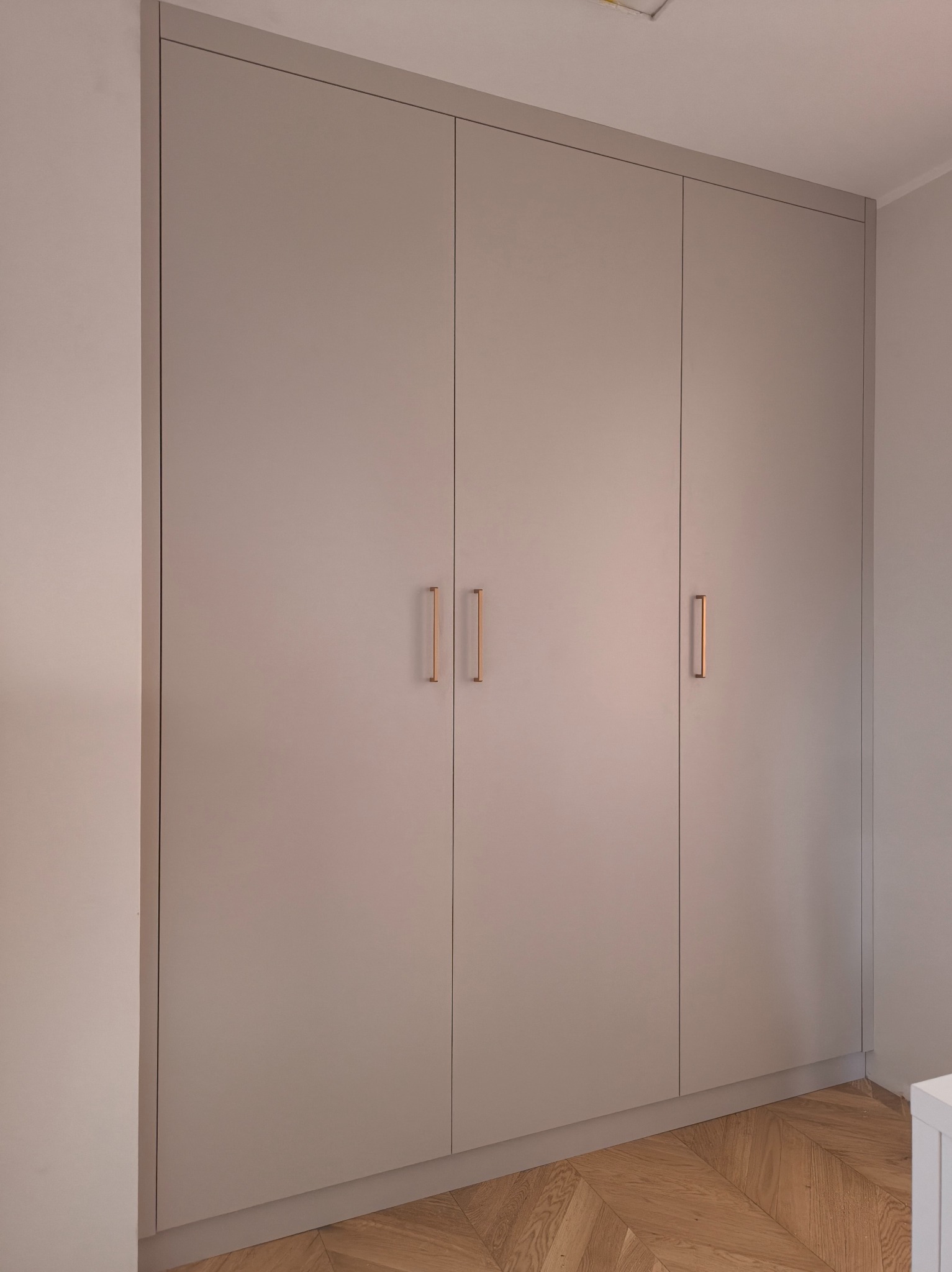 Elegancka, beżowa szafa wnękowa z trzema drzwiami i minimalistycznymi, pionowymi uchwytami w kolorze miedzi, na tle parkietu ułożonego w jodełkę.