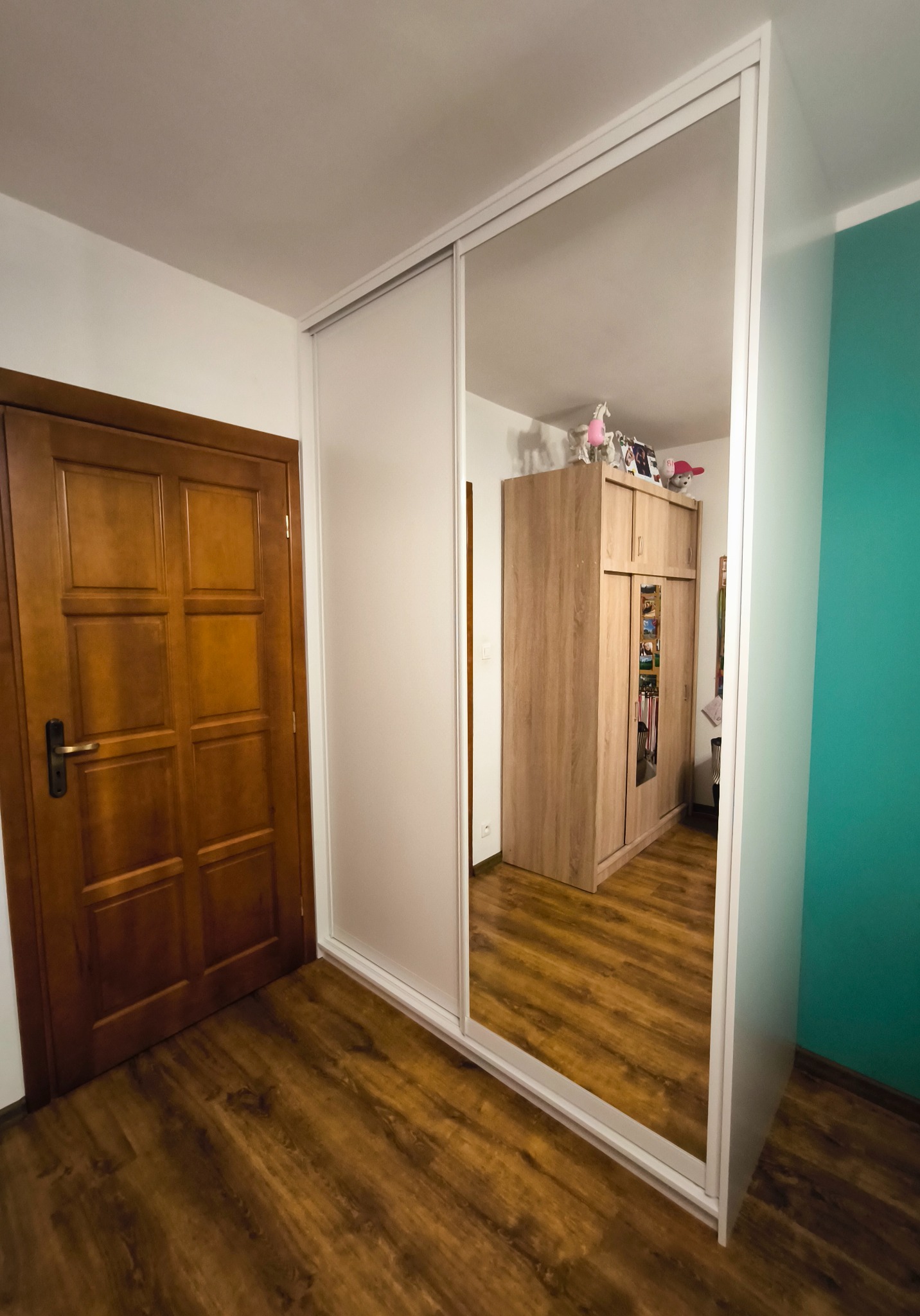 Biała szafa wnękowa z lustrzanymi drzwiami przesuwnymi, odbijająca wnętrze pokoju z drewnianą podłogą i szafą. Widok z perspektywy podłogi.