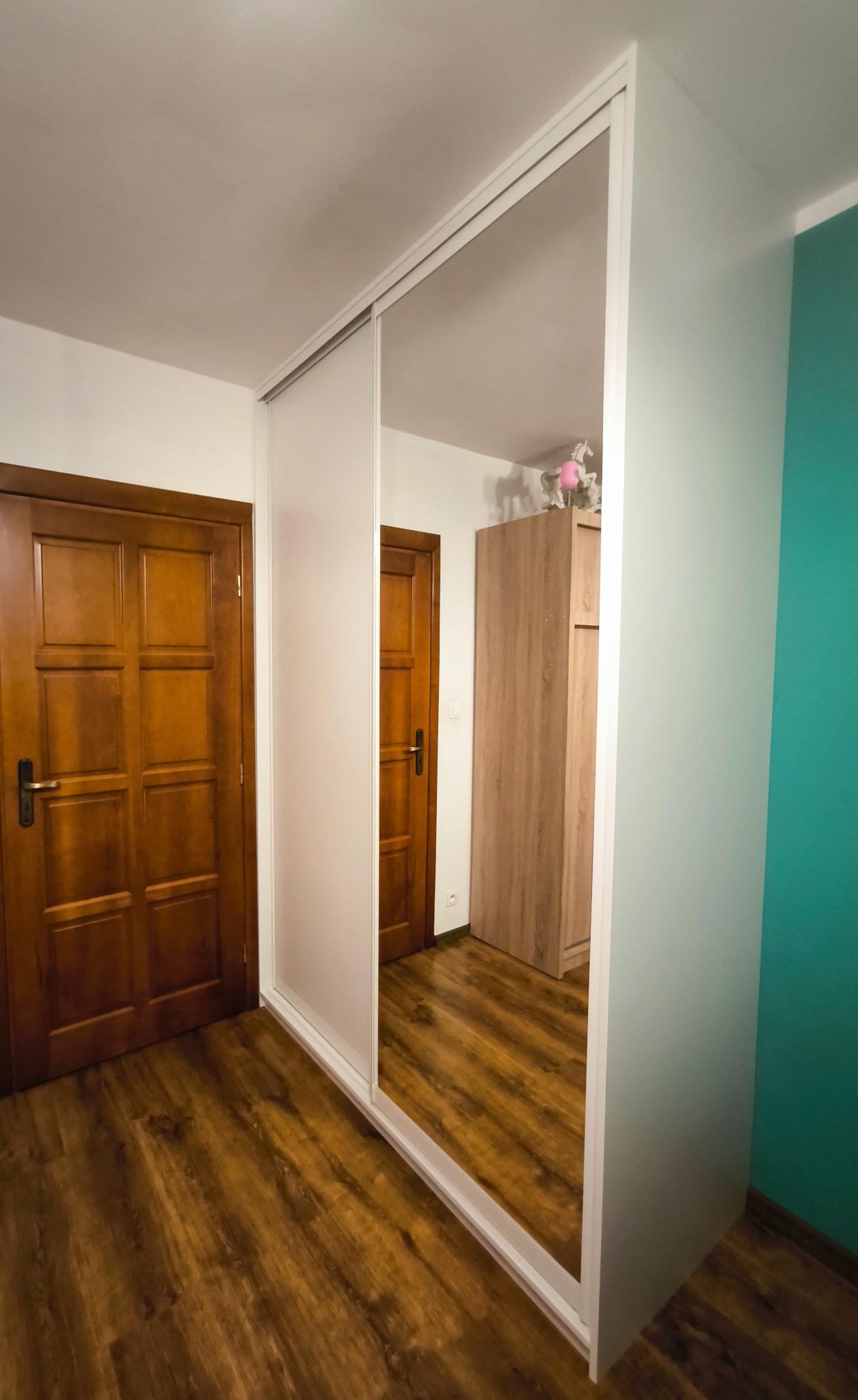 Biała szafa przesuwna z lustrem w przedpokoju. Widoczne drewniane drzwi i podłoga. W lustrze odbija się szafa i zabawka jednorożca.