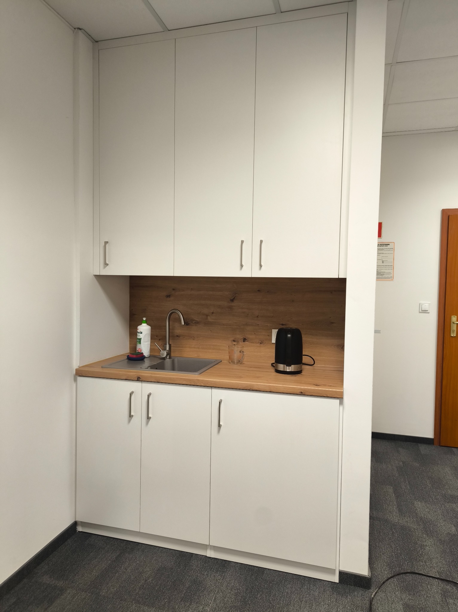 Biała zabudowa kuchenna z drewnianym blatem i zlewozmywakiem w biurze. Na blacie czajnik elektryczny i płyn do naczyń. Minimalistyczny design, szara wykładzina.