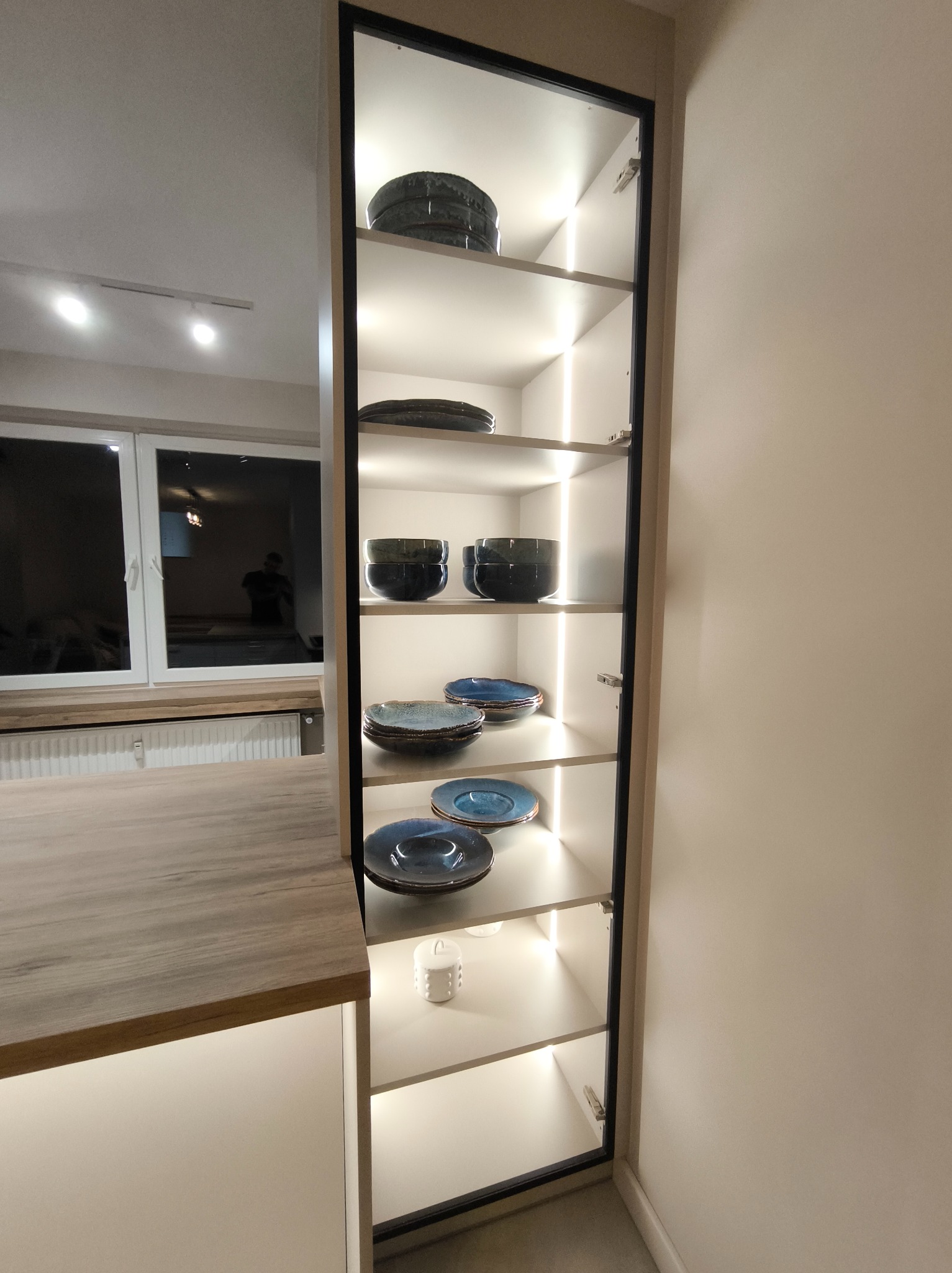 Pionowa szafka kuchenna z podświetlanymi półkami, na których ułożono naczynia. Widoczny fragment blatu i okno w tle. Nowoczesny design, ciepłe oświetlenie.