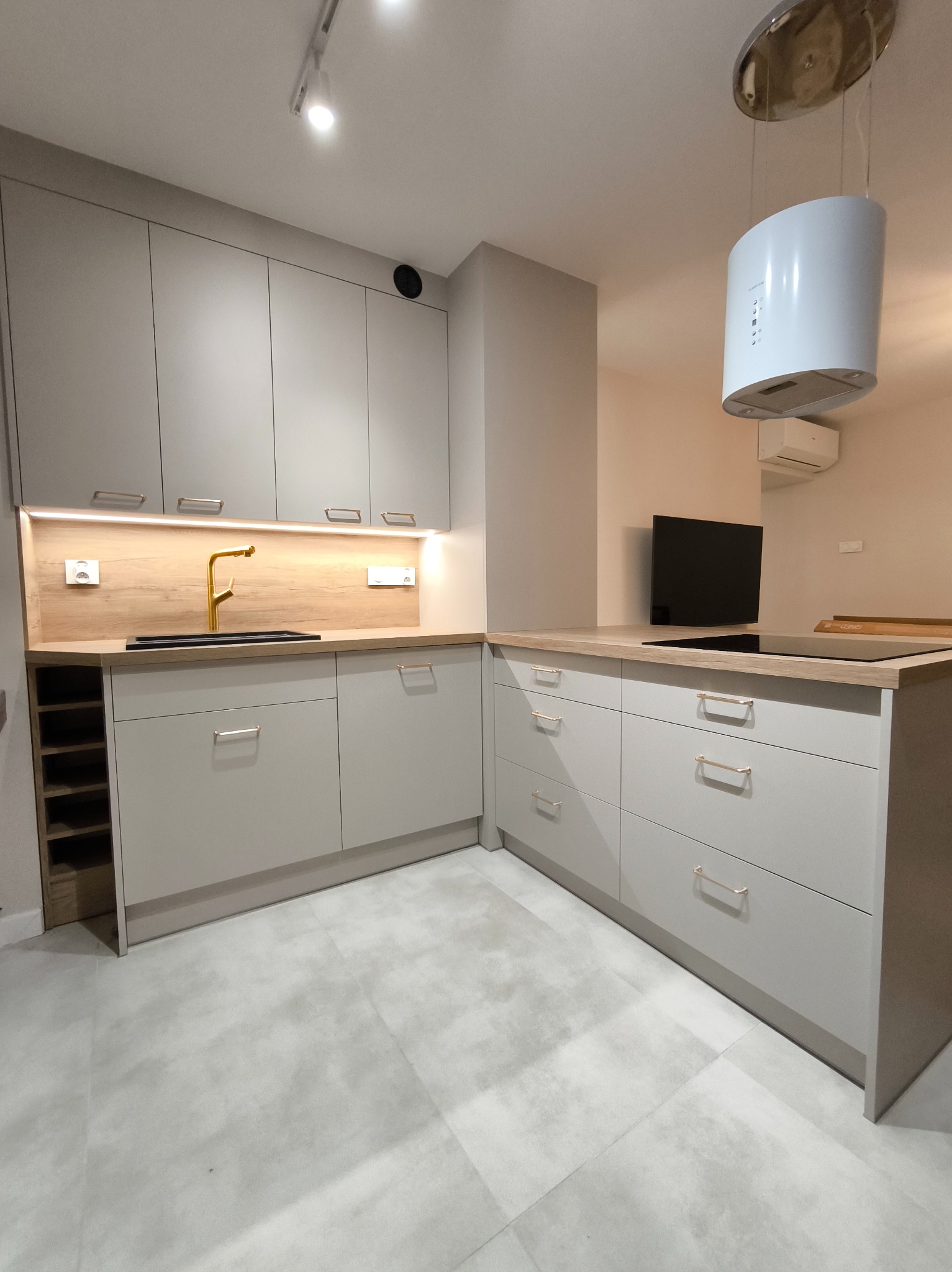 Jasnoszara kuchnia z drewnianym blatem i złotą baterią. Widoczny okap cylindryczny i płyta indukcyjna. Minimalistyczny design, nowoczesne oświetlenie podszafkowe.