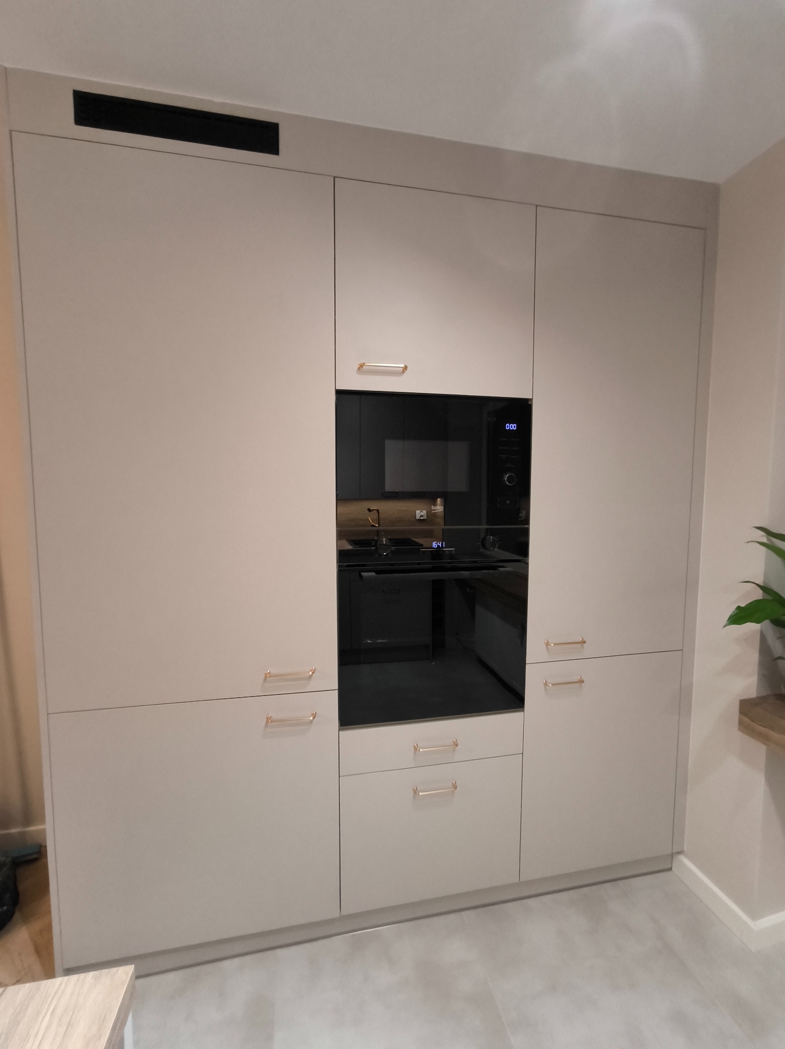 Jasna zabudowa kuchenna z piekarnikiem i mikrofalówką w czarnych obudowach. Minimalistyczne złote uchwyty. Widoczne odbicie wnętrza kuchni w szybie piekarnika.