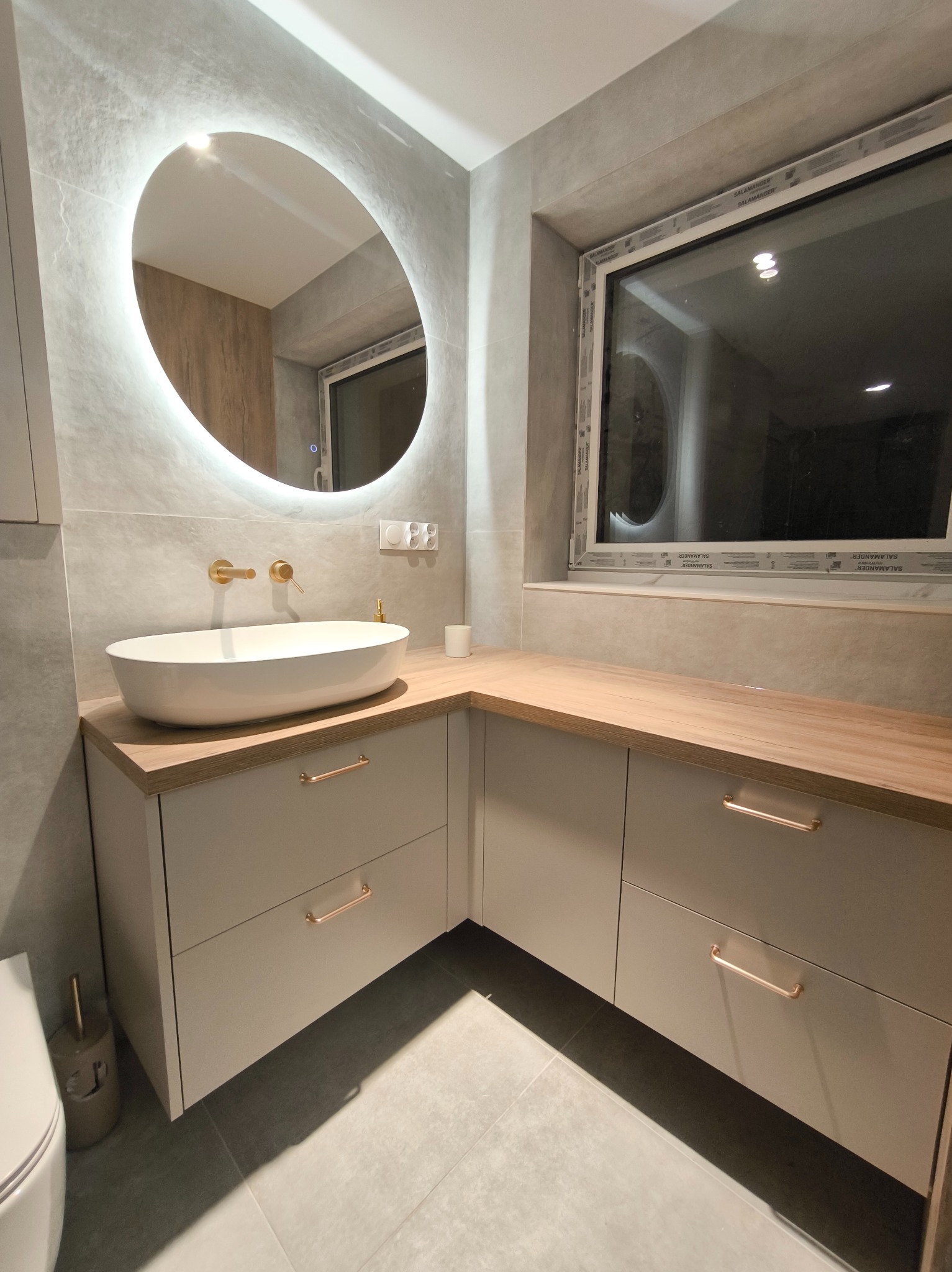 Nowoczesna łazienka z owalną umywalką nablatową, złotą armaturą i podświetlanym lustrem. Jasne szafki z drewnianym blatem i złotymi uchwytami.