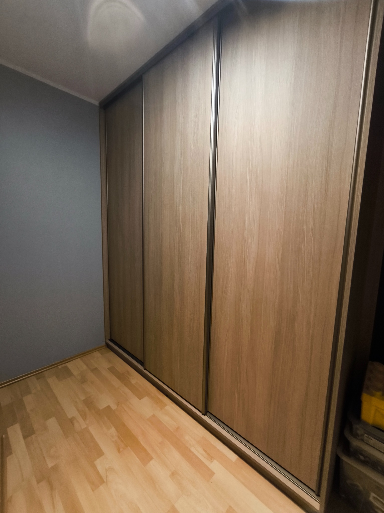 Trzydrzwiowa szafa wnękowa z przesuwnymi drzwiami w odcieniach drewna, z metalowymi uchwytami, na tle szarej ściany i jasnej drewnianej podłogi.