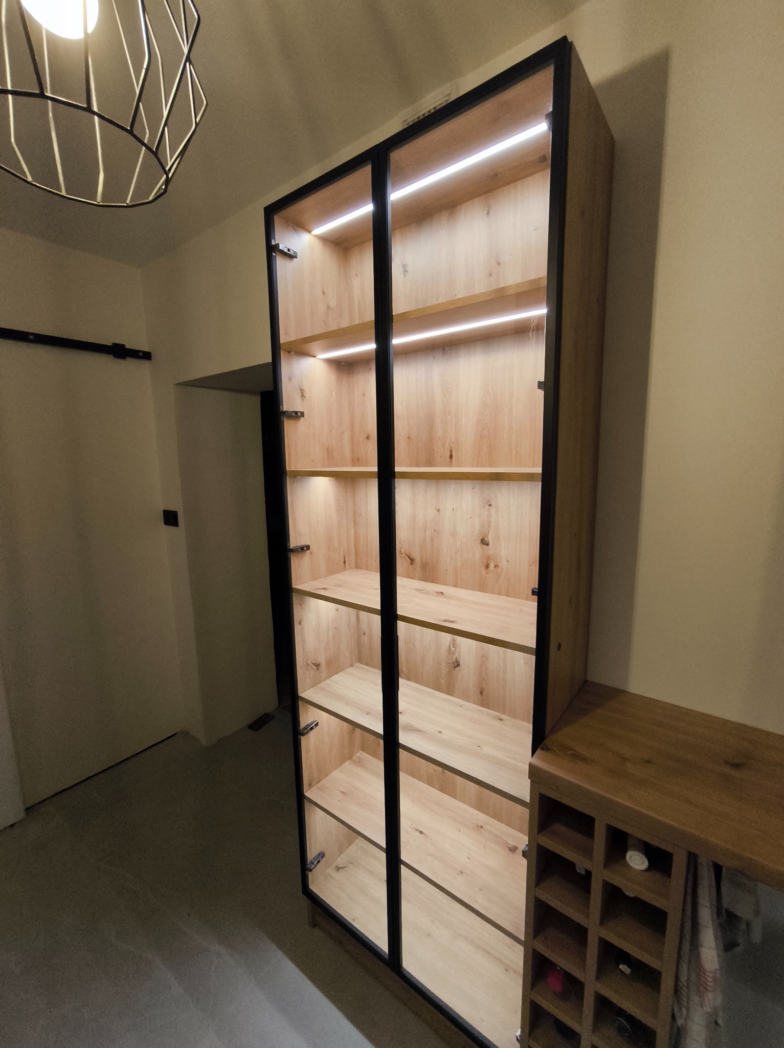 Nowoczesna, wysoka szafa z przeszklonymi drzwiami i oświetleniem LED, wykonana z drewna w ciepłym odcieniu. Obok szafki widoczny regał na wino, całość w neutralnej tonacji.