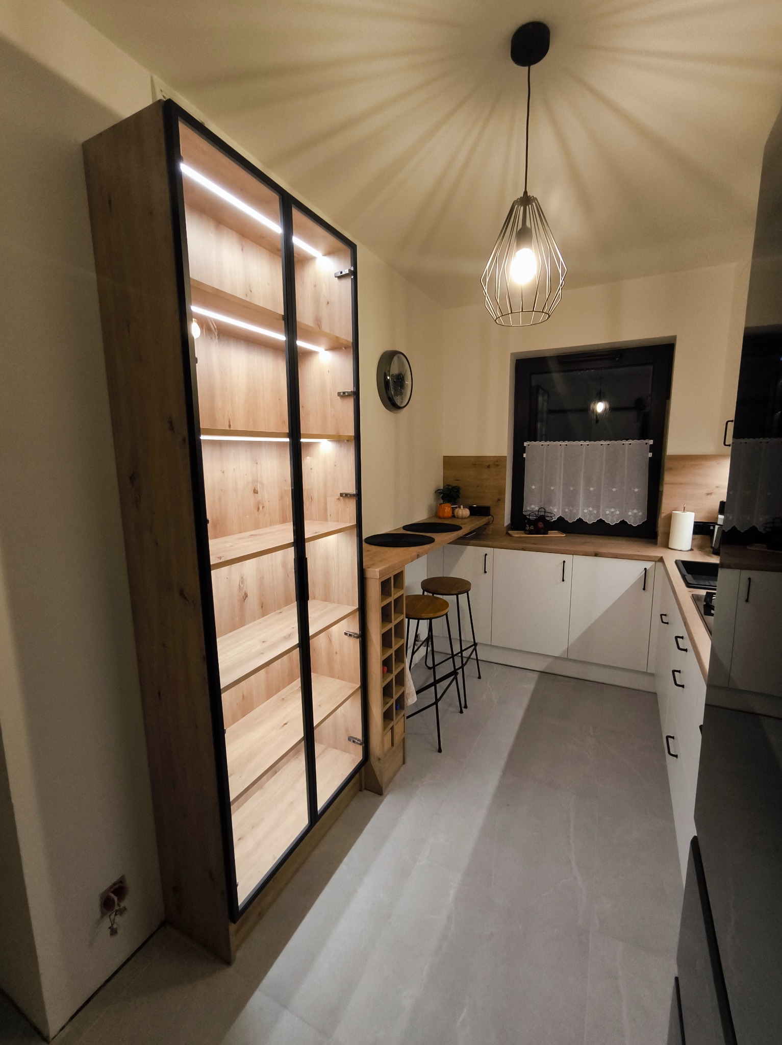 Nowoczesna kuchnia z wysoką, podświetlaną szafą z przeszklonymi drzwiami i drewnianymi półkami, widok na blat z hokerami i okno z firanką.