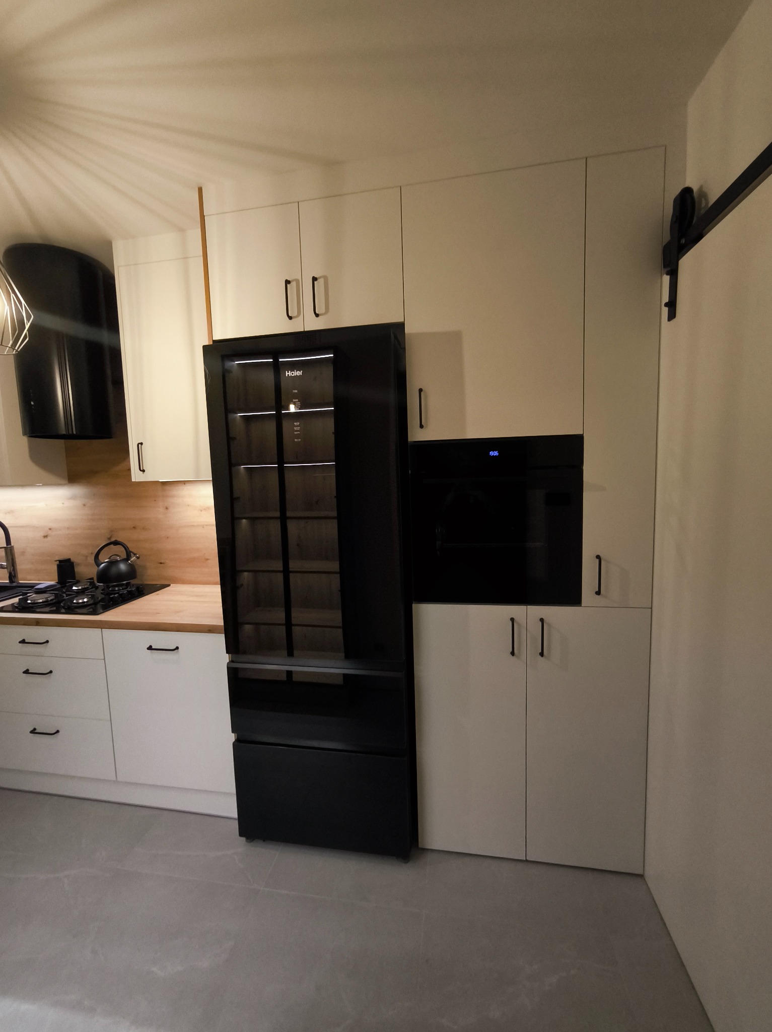 Nowoczesna kuchnia z białymi szafkami, czarnym piekarnikiem i lodówką Haier. Blat drewniany, płyta gazowa i czarny okap. Minimalistyczny design.