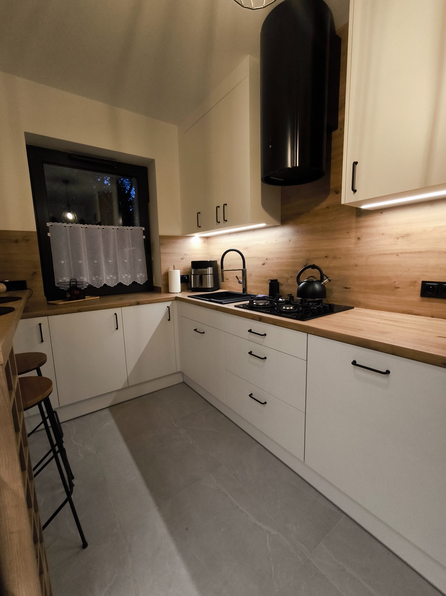 Nowoczesna kuchnia z białymi szafkami, drewnianym blatem i czarnymi akcentami, w tym okapem i armaturą. Uwagę przykuwa oświetlenie LED pod szafkami.