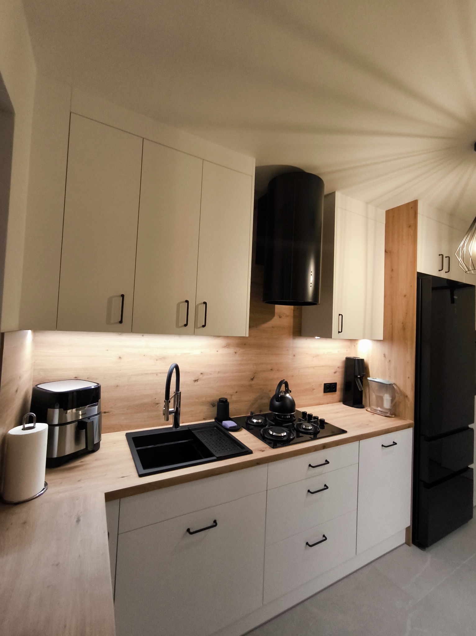 Nowoczesna kuchnia z białymi szafkami, czarnym zlewem i sprzętami AGD. Blat drewnopodobny, oświetlenie LED pod szafkami. Minimalistyczny design z czarnymi uchwytami.