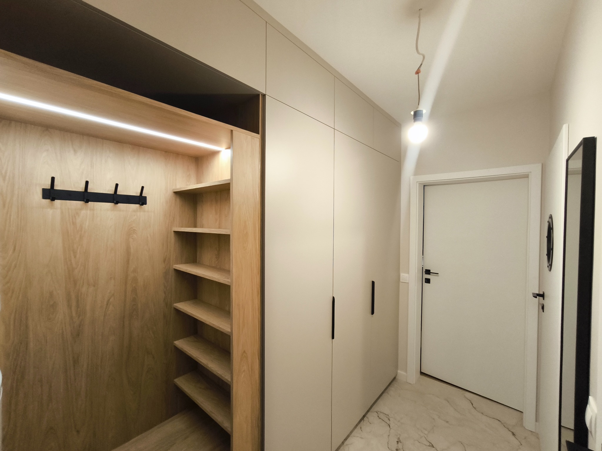 Jasna szafa wnękowa z drewnianym regałem i oświetleniem LED w przedpokoju. Minimalistyczny design, czarne uchwyty, marmurowa podłoga. Widok na drzwi.