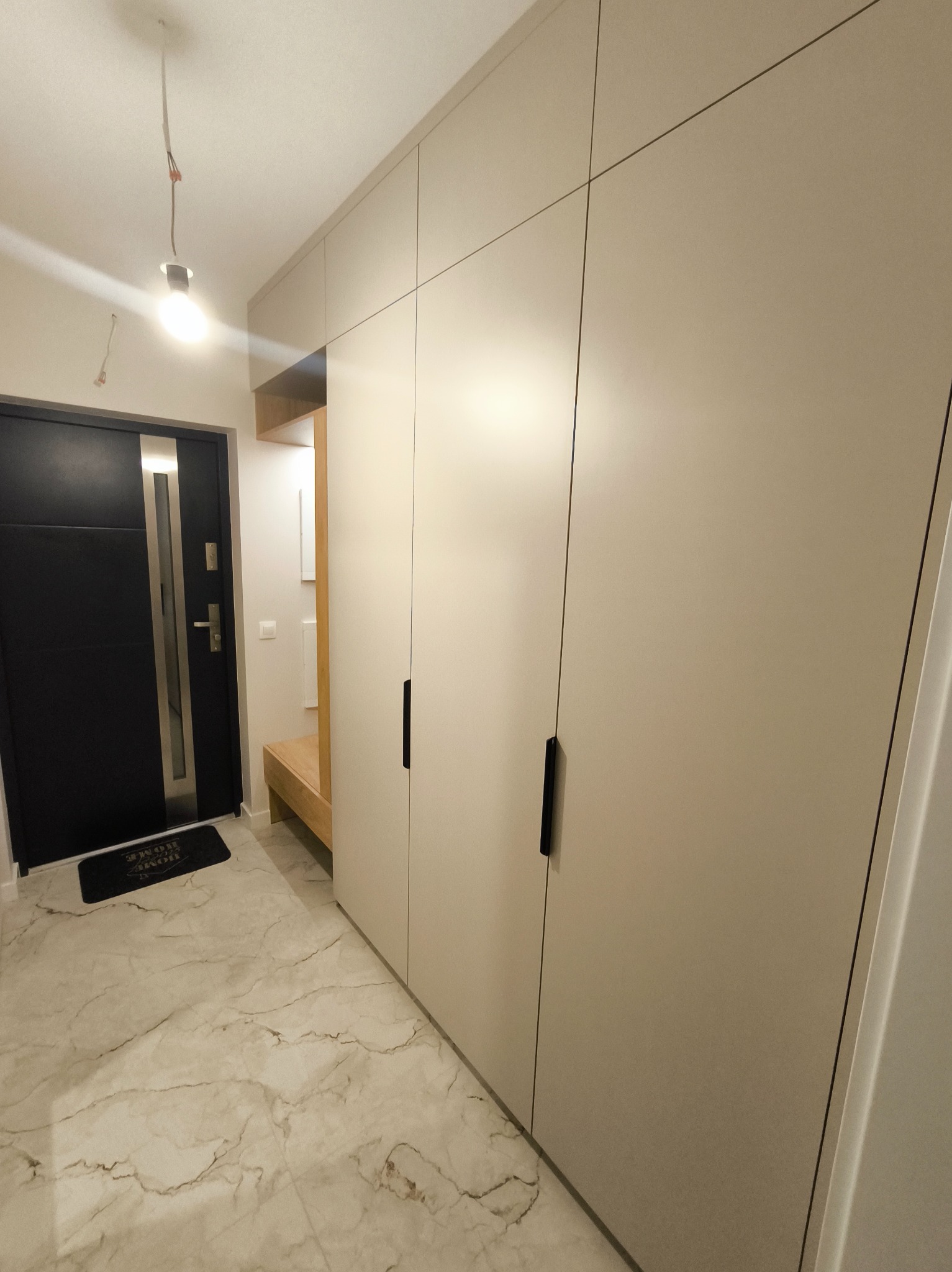 Jasna szafa wnękowa z czarnymi uchwytami, w przedpokoju z marmurową podłogą i czarnymi drzwiami wejściowymi. Widoczna żarówka bez klosza.