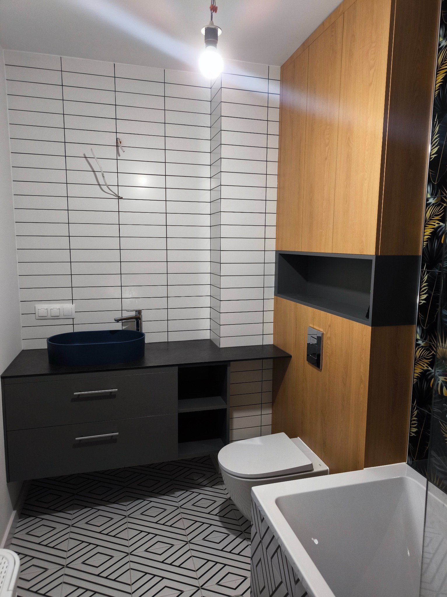 Nowoczesna łazienka z drewnianą szafą w zabudowie, umywalką w kolorze granatowym i geometryczną podłogą. Widoczne białe płytki na ścianie i fragment wanny.