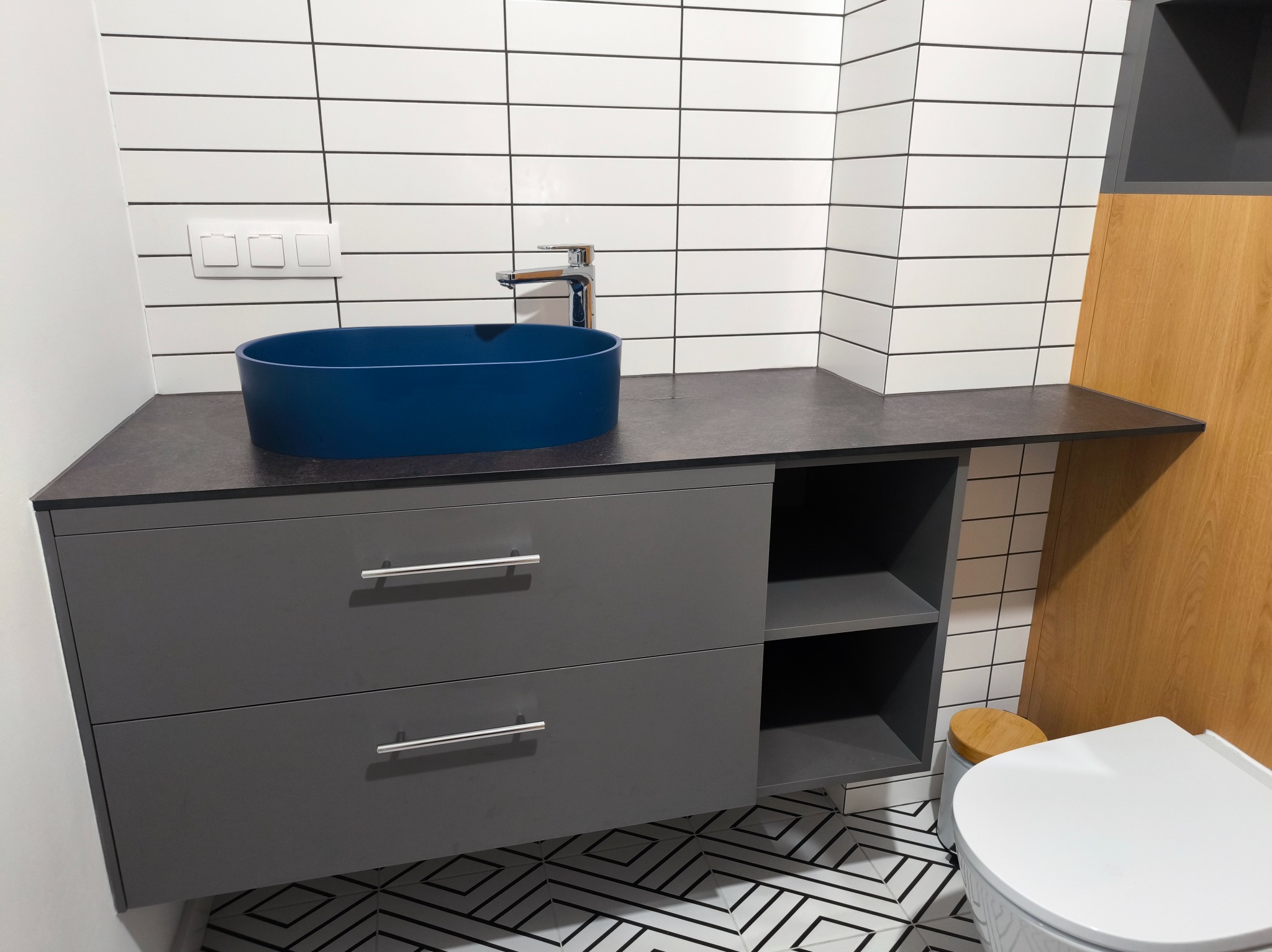 Szara szafka łazienkowa z ciemnym blatem, na którym stoi okrągła, niebieska umywalka. W tle białe płytki z czarnymi fugami i fragment toalety.