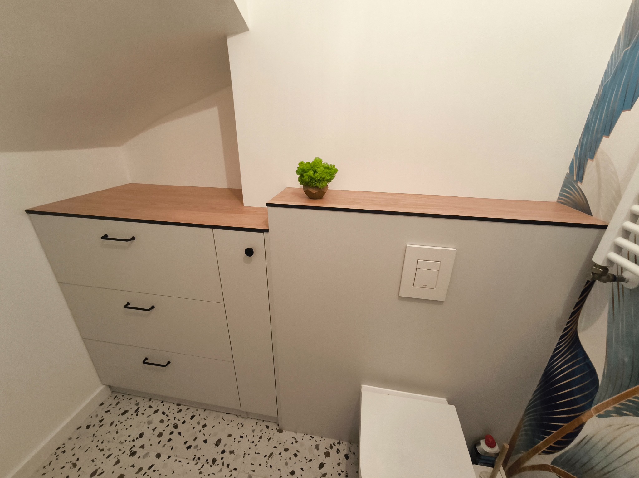 Biała szafka z trzema szufladami i drewnianym blatem w łazience na poddaszu. Na blacie dekoracyjna roślina. Podłoga w stylu lastryko.