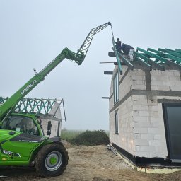 Roboty Ziemne Patryk Makarski - Budowa domu z bloczków: montaż więźby dachowej z użyciem podnośnika teleskopowego Merlo. Widoczny surowy stan budynku i pracownicy na dachu.