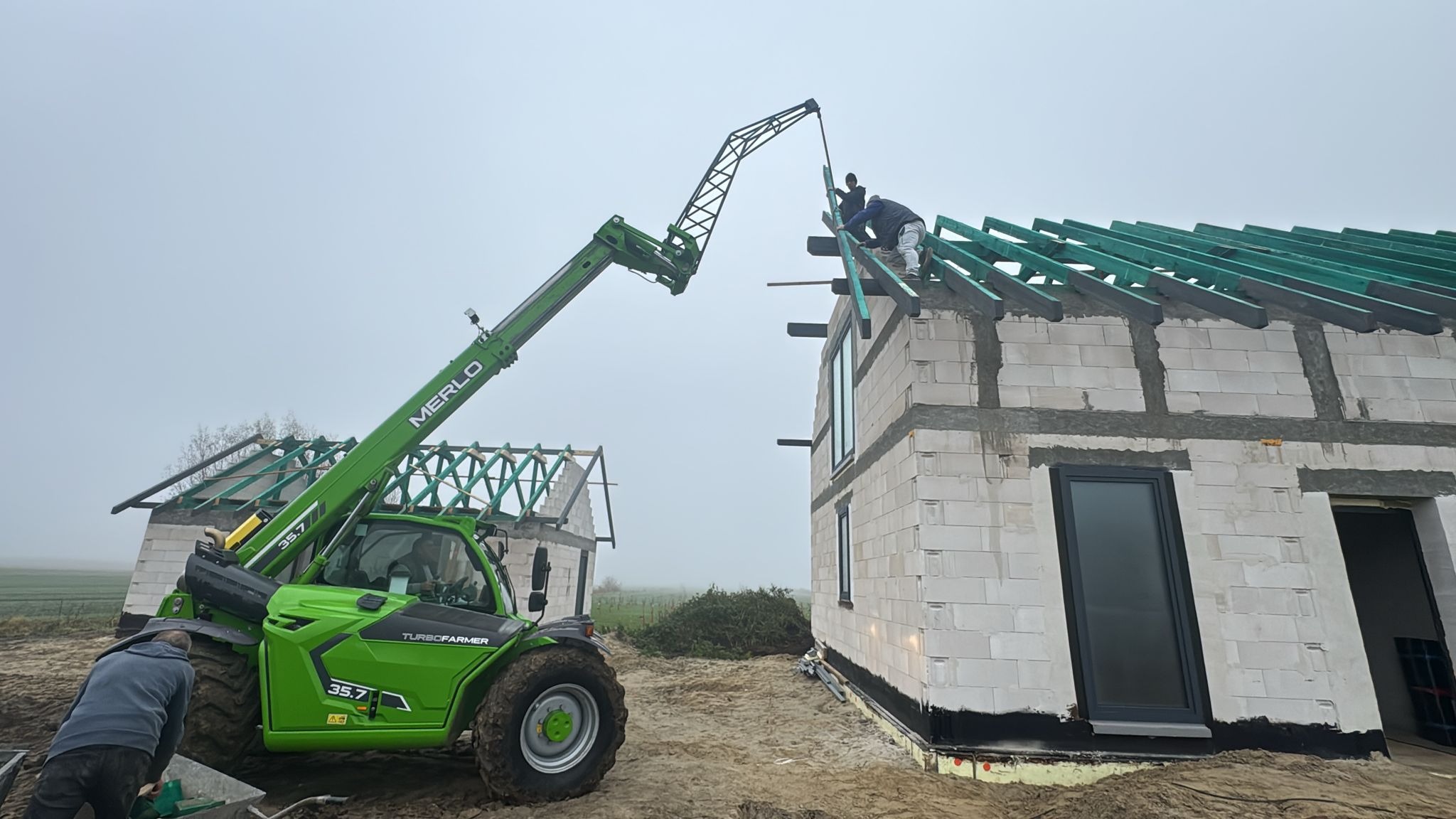 Budowa domu z bloczków: montaż więźby dachowej z użyciem podnośnika teleskopowego Merlo. Widoczny surowy stan budynku i pracownicy na dachu.