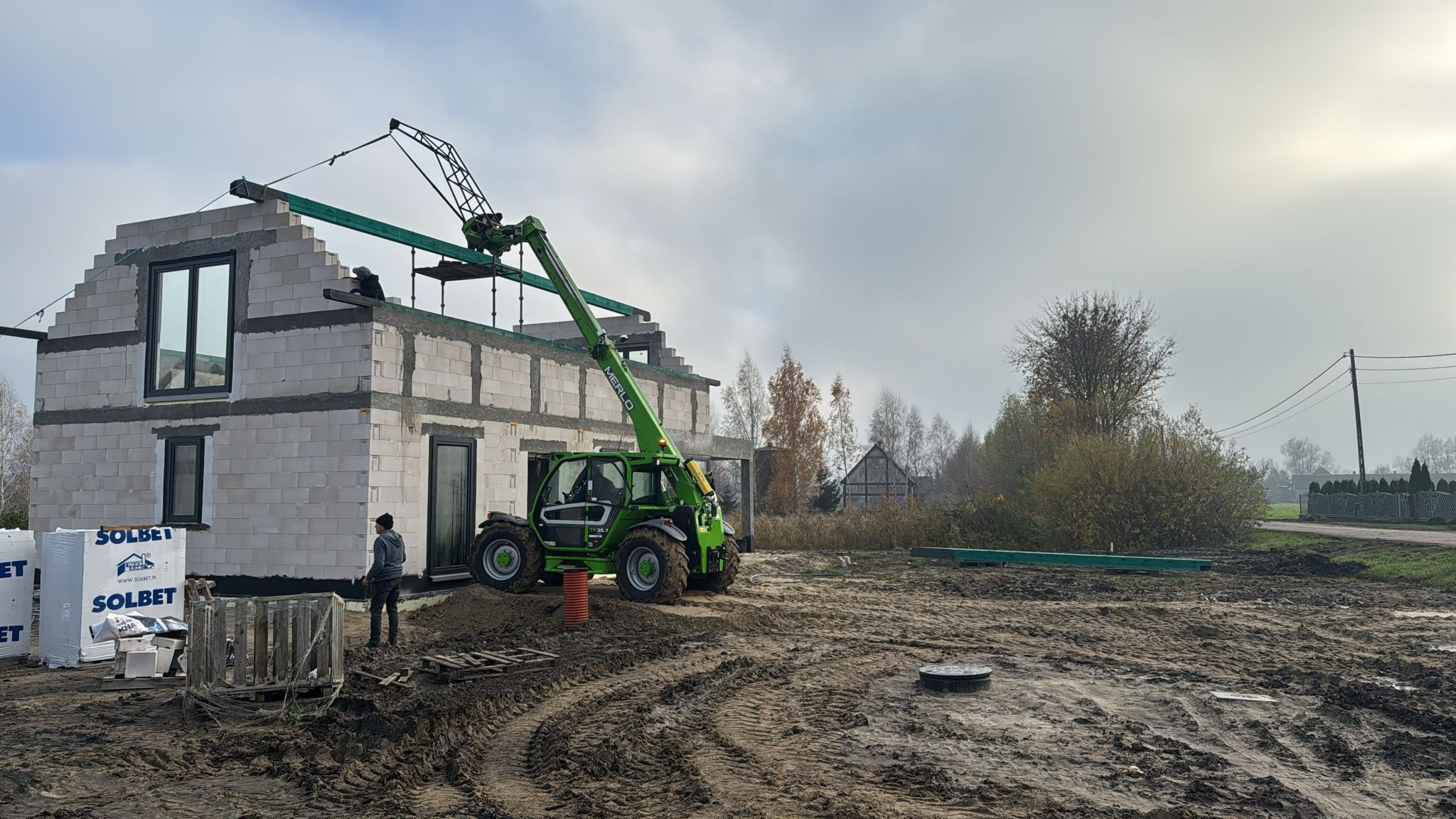 Budowa domu z betonu komórkowego w trakcie: zielony podnośnik teleskopowy Merlo podnosi belkę, pracownik na dachu, palety z bloczkami Solbet obok. Błotniste podłoże.