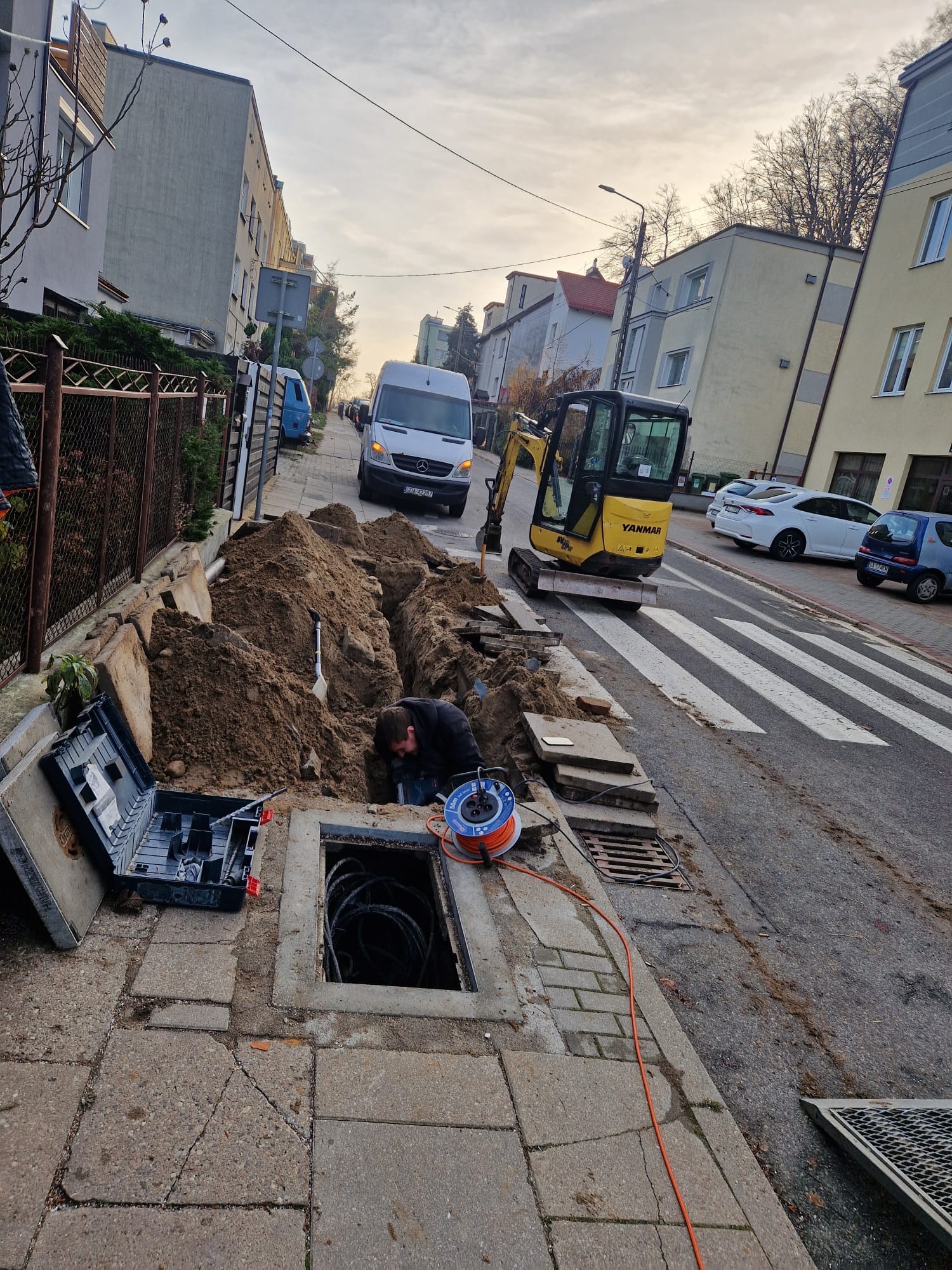 Prace ziemne na ulicy: wykop z kablami, mini koparka Yanmar, narzędzia i pracownik w czarnej kurtce. W tle budynki mieszkalne i zaparkowane samochody.