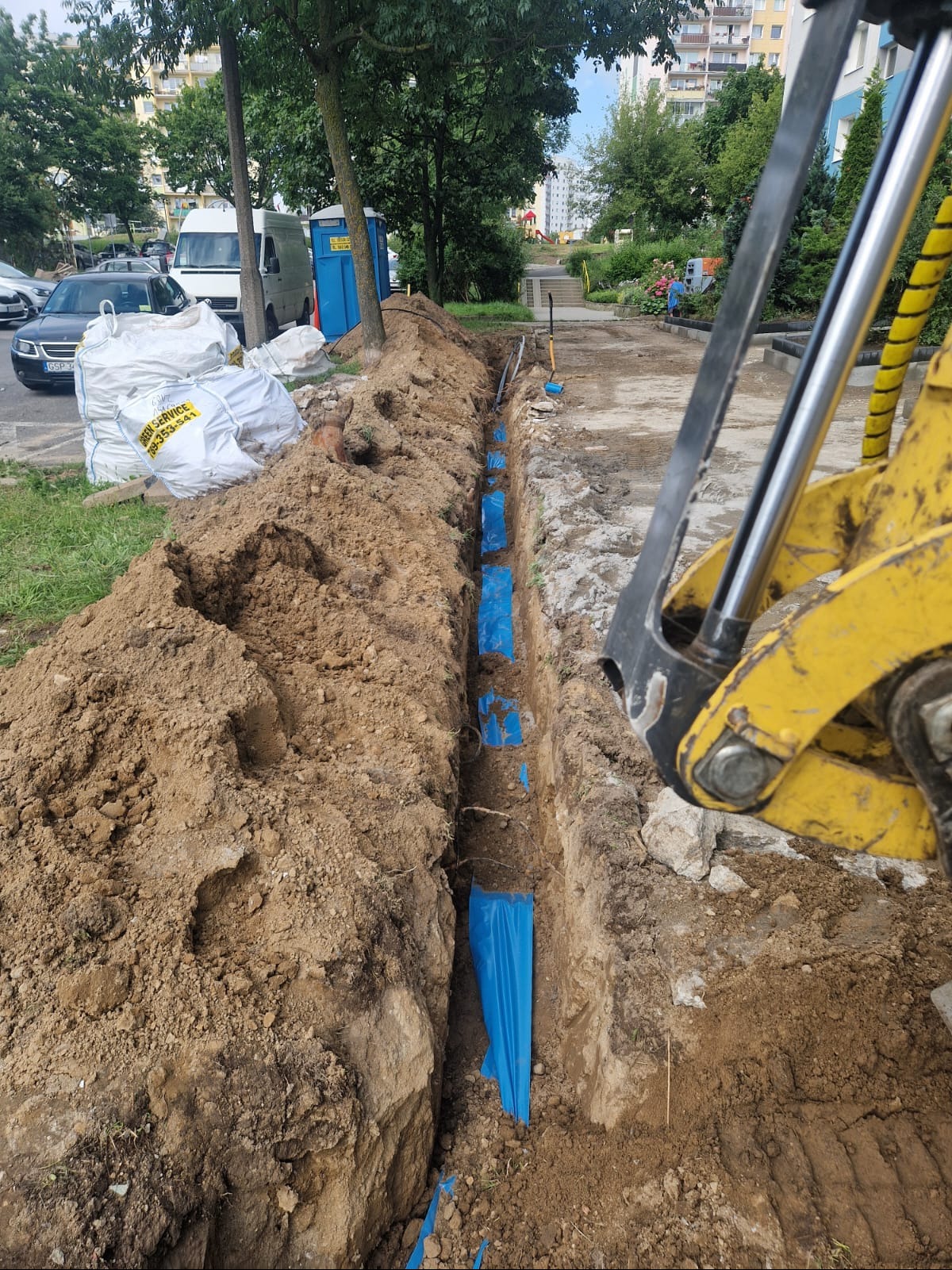 Wykopany rów z niebieską folią zabezpieczającą, widoczny fragment koparki, w tle budynki mieszkalne i worki z ziemią. Prace ziemne w przestrzeni miejskiej.