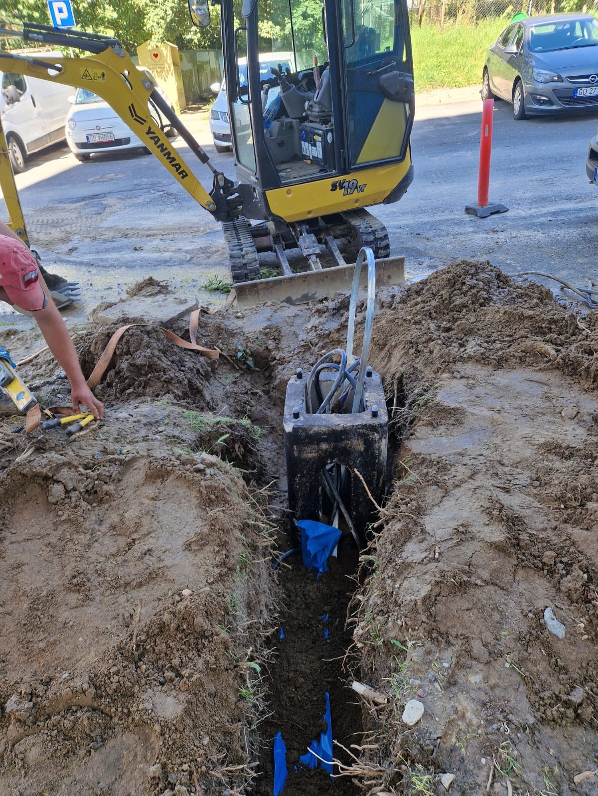 Żółta mini koparka Yanmar przy wykopie z kablem w czarnej skrzynce. Widoczne narzędzia i ręka pracownika. Prace ziemne w trakcie realizacji, z bliska.