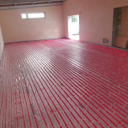 INSTAL-ART S.C. T. KLOC, S. DUDEK. - Rozłożona instalacja ogrzewania podłogowego z czerwonymi rurkami na siatce w surowym wnętrzu, widoczne okno i otwór drzwiowy.