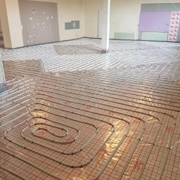 INSTAL-ART S.C. T. KLOC, S. DUDEK. - Rozłożone rury ogrzewania podłogowego na folii z czerwoną siatką, widoczny betonowy słup w surowym pomieszczeniu w trakcie budowy.