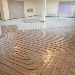 Rozłożone rury ogrzewania podłogowego na folii z czerwoną siatką, widoczny betonowy słup w surowym pomieszczeniu w trakcie budowy.