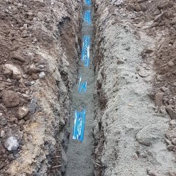 INSTAL-ART S.C. T. KLOC, S. DUDEK. - Wykop z ułożonymi rurami w niebieskiej folii, zasypywany piaskiem, widoczni pracownicy i koparka w tle.