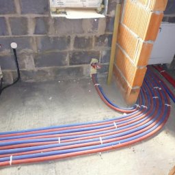 INSTAL-ART S.C. T. KLOC, S. DUDEK. - Wiązka czerwono-niebieskich rur instalacji wodnej lub grzewczej prowadzona w surowym stanie podłogi, mocowana do ściany z pustaków, widoczne elementy instalacji elektrycznej.