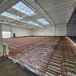 INSTAL-ART S.C. T. KLOC, S. DUDEK. - Rozległa hala przemysłowa z widoczną instalacją ogrzewania podłogowego w trakcie montażu, rury grzewcze ułożone na srebrnej folii izolacyjnej, betonowy filar w lewym dolnym rogu, błękitne...