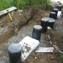 INSTAL-ART S.C. T. KLOC, S. DUDEK. - Wykop z ułożonym drenażem, czarne studzienki rozsączające i żółta łopata oparta o brzeg wykopu.