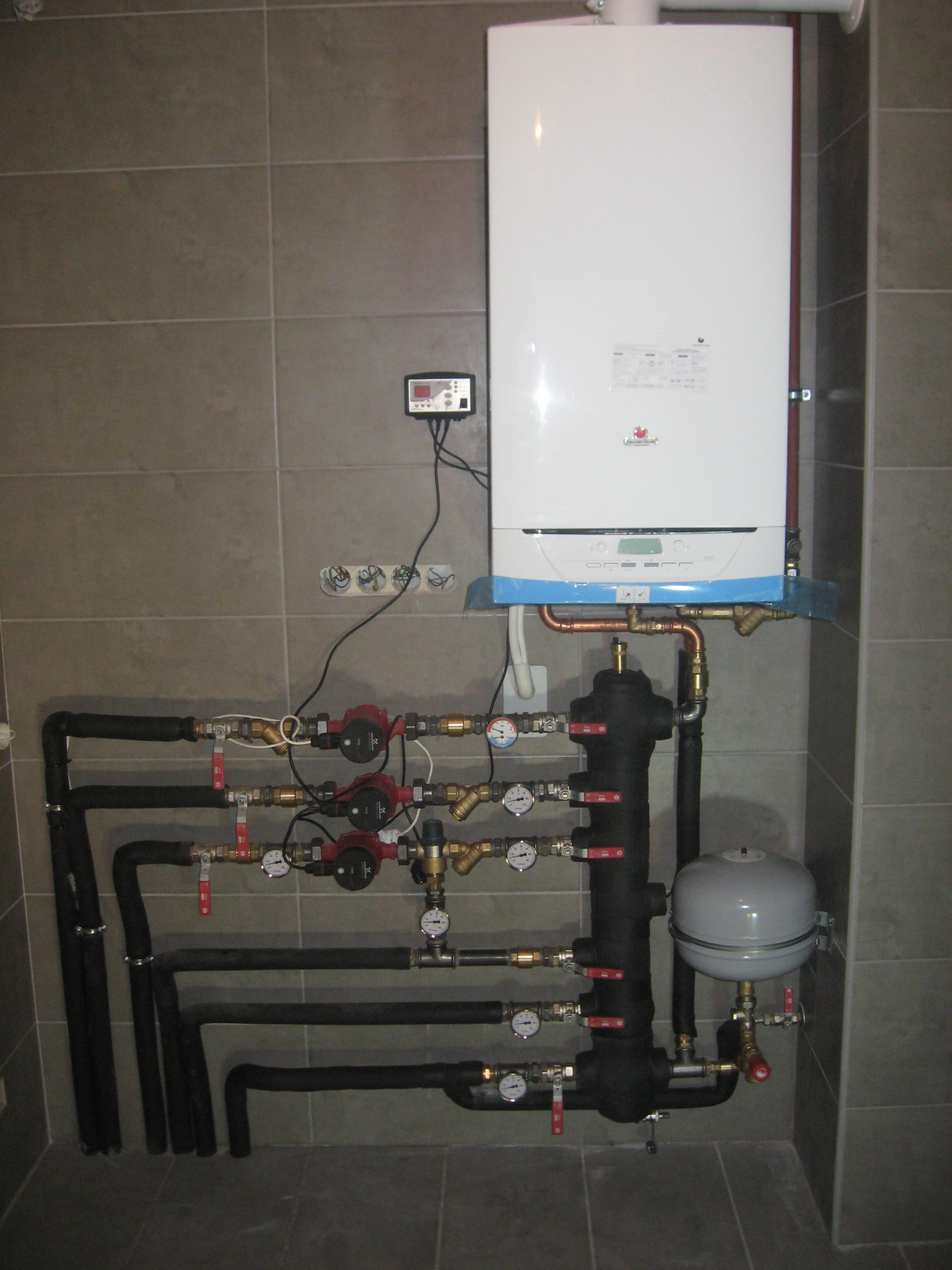 Biały piec gazowy z podłączeniami hydraulicznymi, czarne rury izolowane, manometry i zawory na tle szarych płytek.