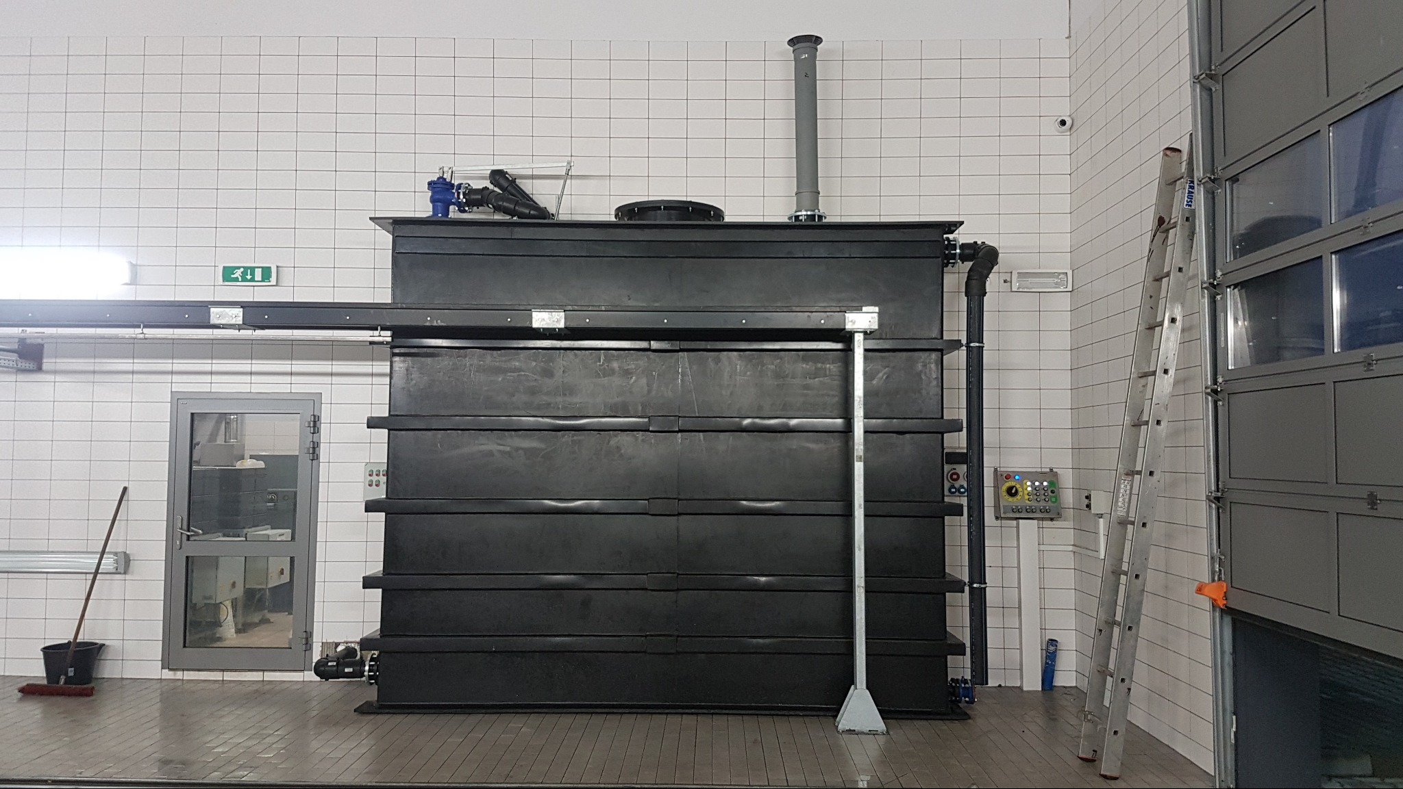 Czarna, segmentowa oczyszczalnia ścieków w pomieszczeniu z białymi kafelkami, widoczna drabina i panel sterowania.