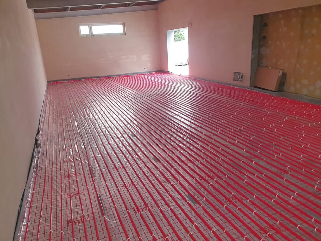 Rozłożona instalacja ogrzewania podłogowego z czerwonymi rurkami na siatce w surowym wnętrzu, widoczne okno i otwór drzwiowy.