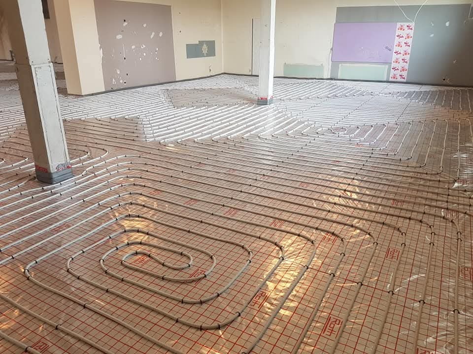 Rozłożone rury ogrzewania podłogowego na folii z czerwoną siatką, widoczny betonowy słup w surowym pomieszczeniu w trakcie budowy.