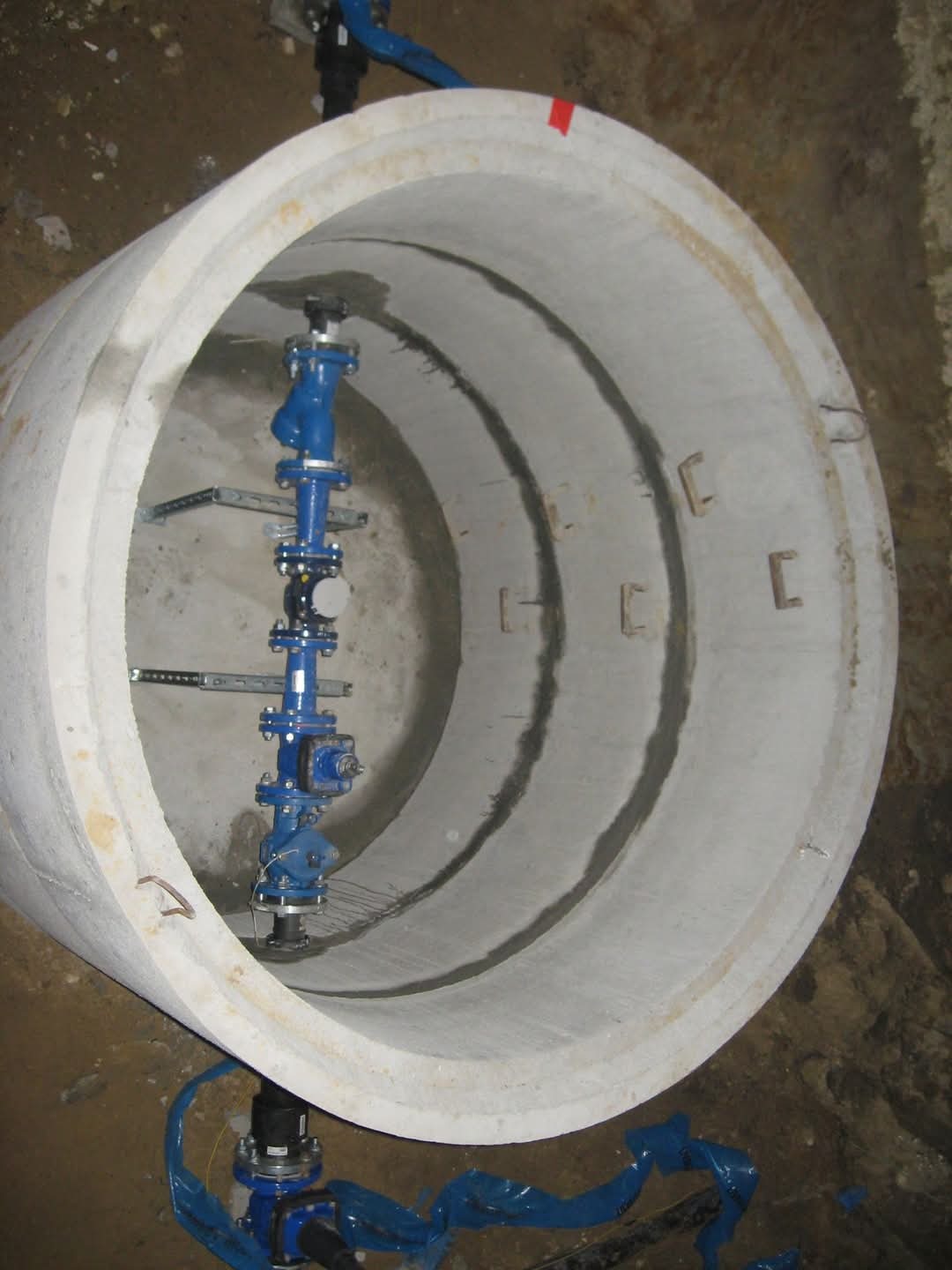 Wnętrze betonowej rury kanalizacyjnej z zamontowaną armaturą wodną w kolorze niebieskim, widoczne elementy mocujące i uszczelnienia.
