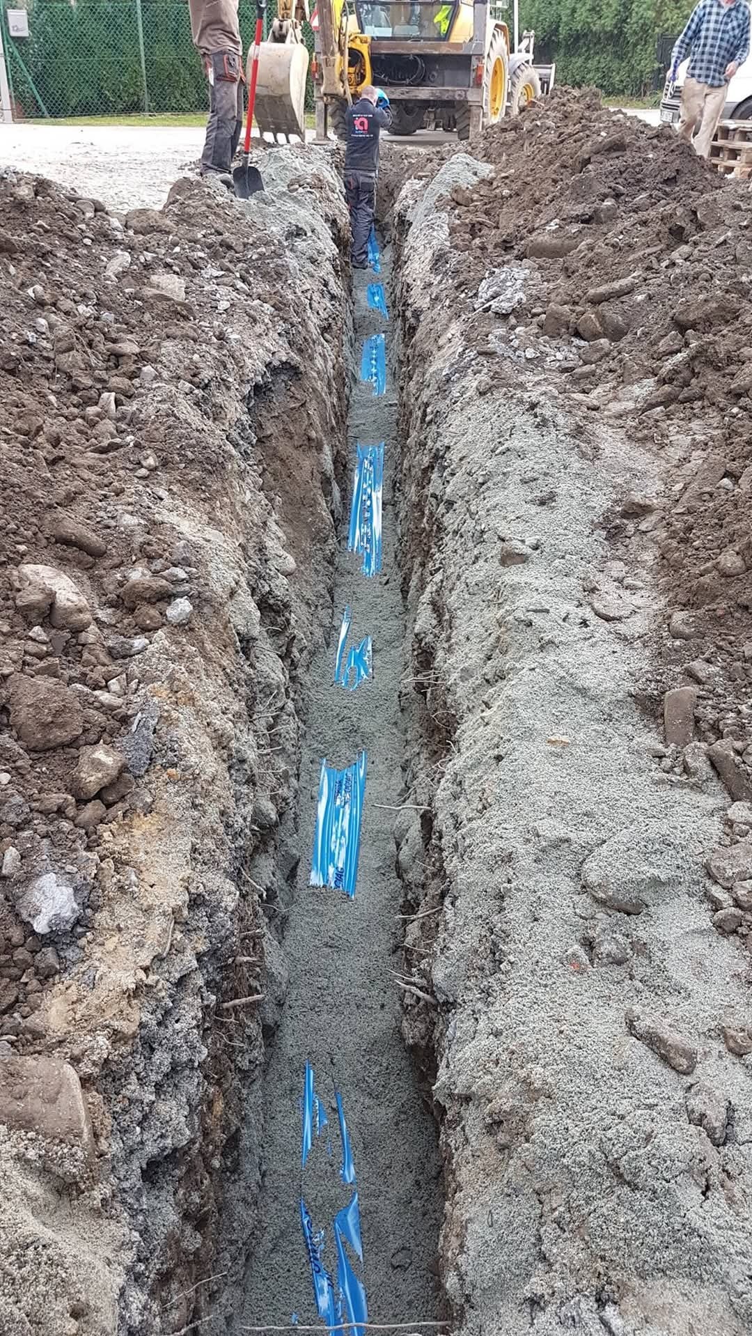 Wykop z ułożonymi rurami w niebieskiej folii, zasypywany piaskiem, widoczni pracownicy i koparka w tle.