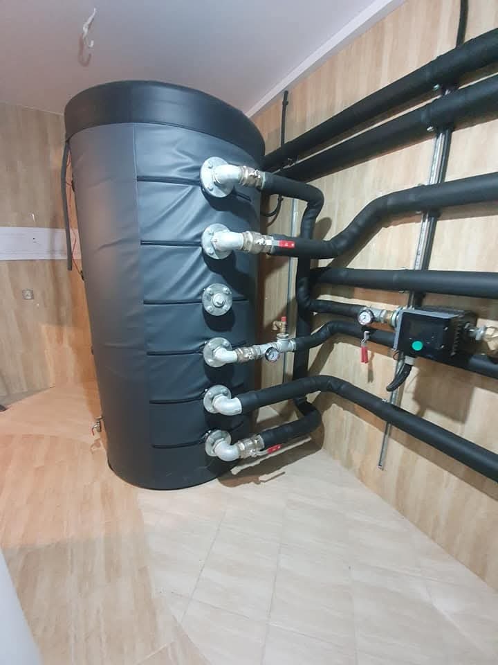 Czarny, izolowany zasobnik ciepłej wody użytkowej z podłączonymi rurami w pomieszczeniu z beżowymi płytkami na ścianach i podłodze.