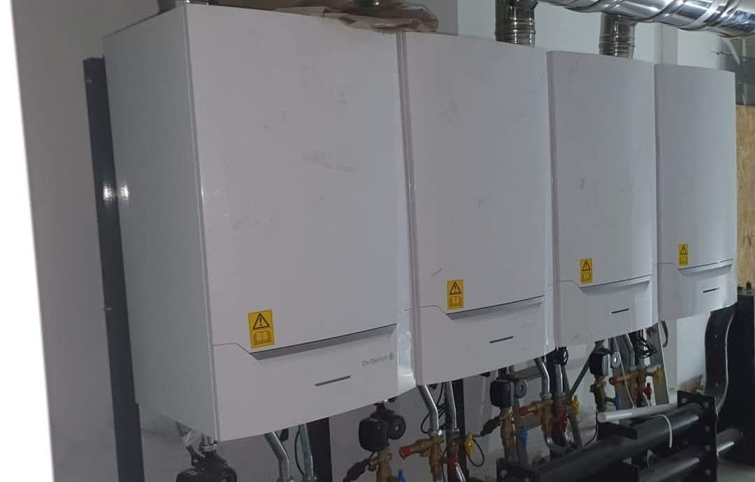 Cztery białe, kondensacyjne piece grzewcze zamontowane szeregowo na ścianie, z widocznymi rurami i zaworami instalacji grzewczej.