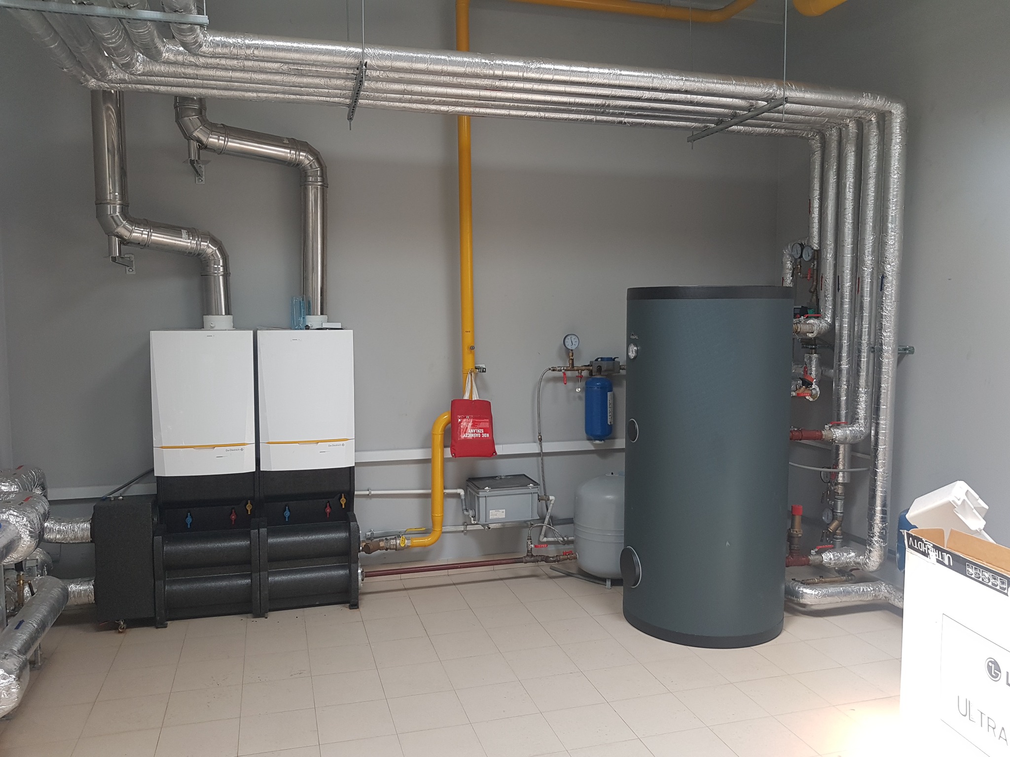Dwa kotły grzewcze De Dietrich z czarnymi modułami, szary pionowy zbiornik, orurowanie w izolacji aluminiowej, żółta rura gazowa i czerwona torba na ścianie w pomieszczeniu z kafelkami na podłodze.