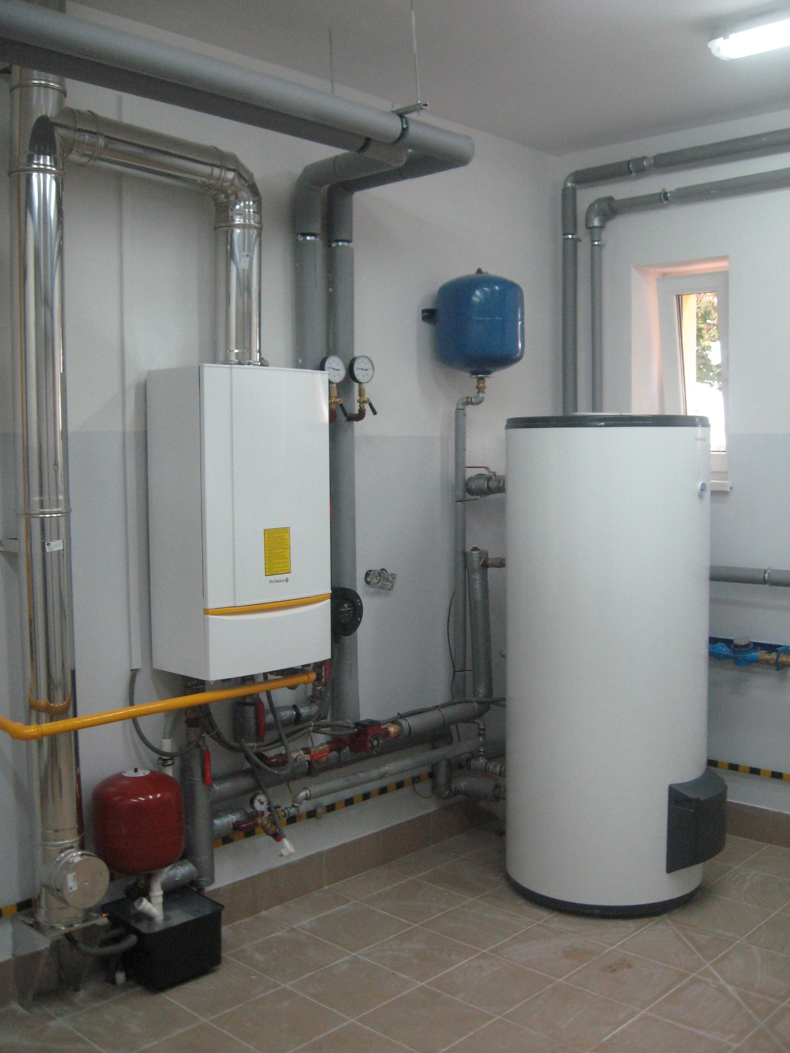 Biały piec CO z żółtymi akcentami, zbiornik akumulacyjny, rury i instalacja w kotłowni, widoczne manometry i zawory, na podłodze płytki ceramiczne.