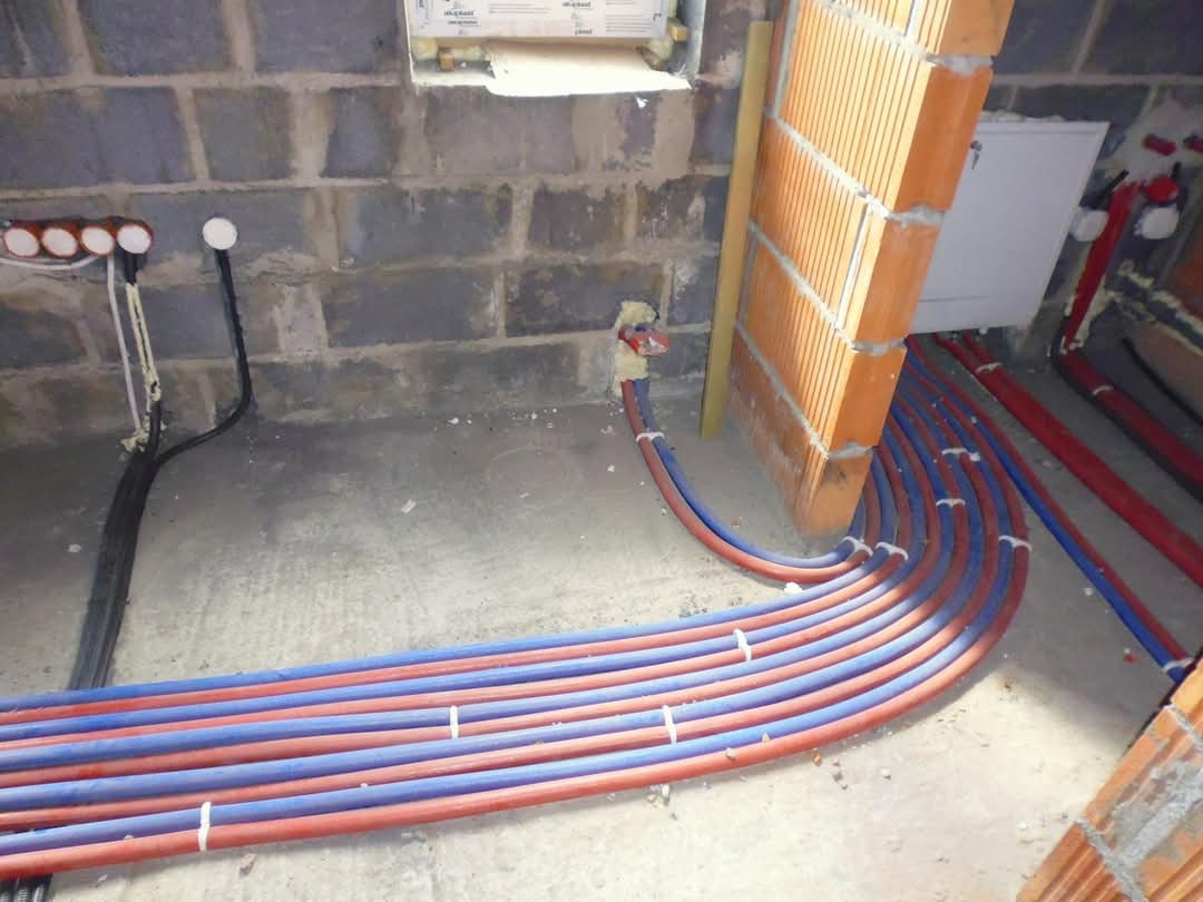 Wiązka czerwono-niebieskich rur instalacji wodnej lub grzewczej prowadzona w surowym stanie podłogi, mocowana do ściany z pustaków, widoczne elementy instalacji elektrycznej.