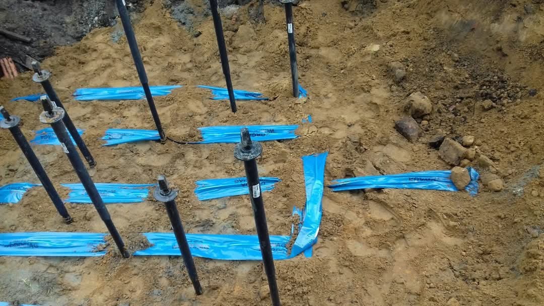 Grunt z wbitymi pionowymi prętami i niebieską folią zabezpieczającą, widoczne ślady prac ziemnych.